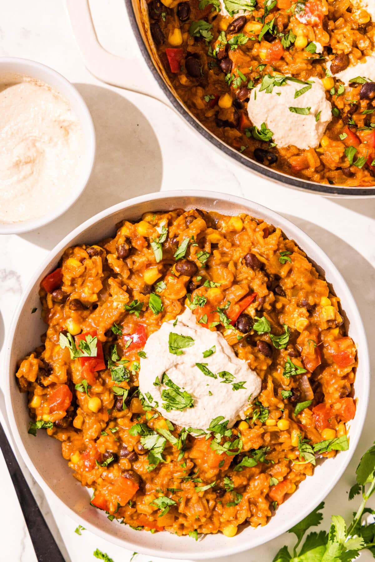 A bowl of a rice and beans dish topped with sour cream and fresh cilantro. Beside the bowl is a smaller bowl of sour cream and a large pot with the remaining rice and beans.