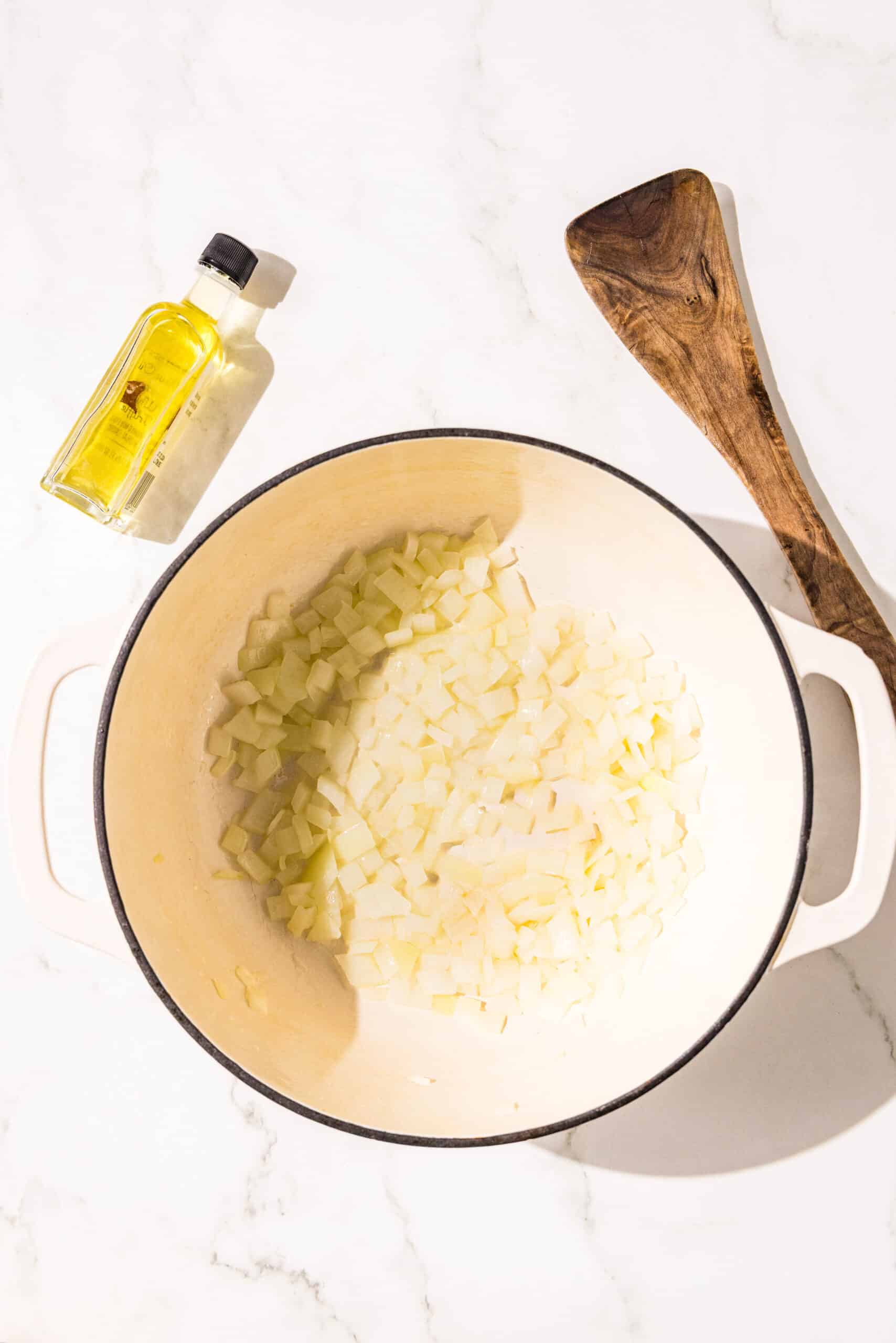 A large pot with oil and diced onions at the bottom. Beside the pot is a wooden spoon and a small bottle of oil.