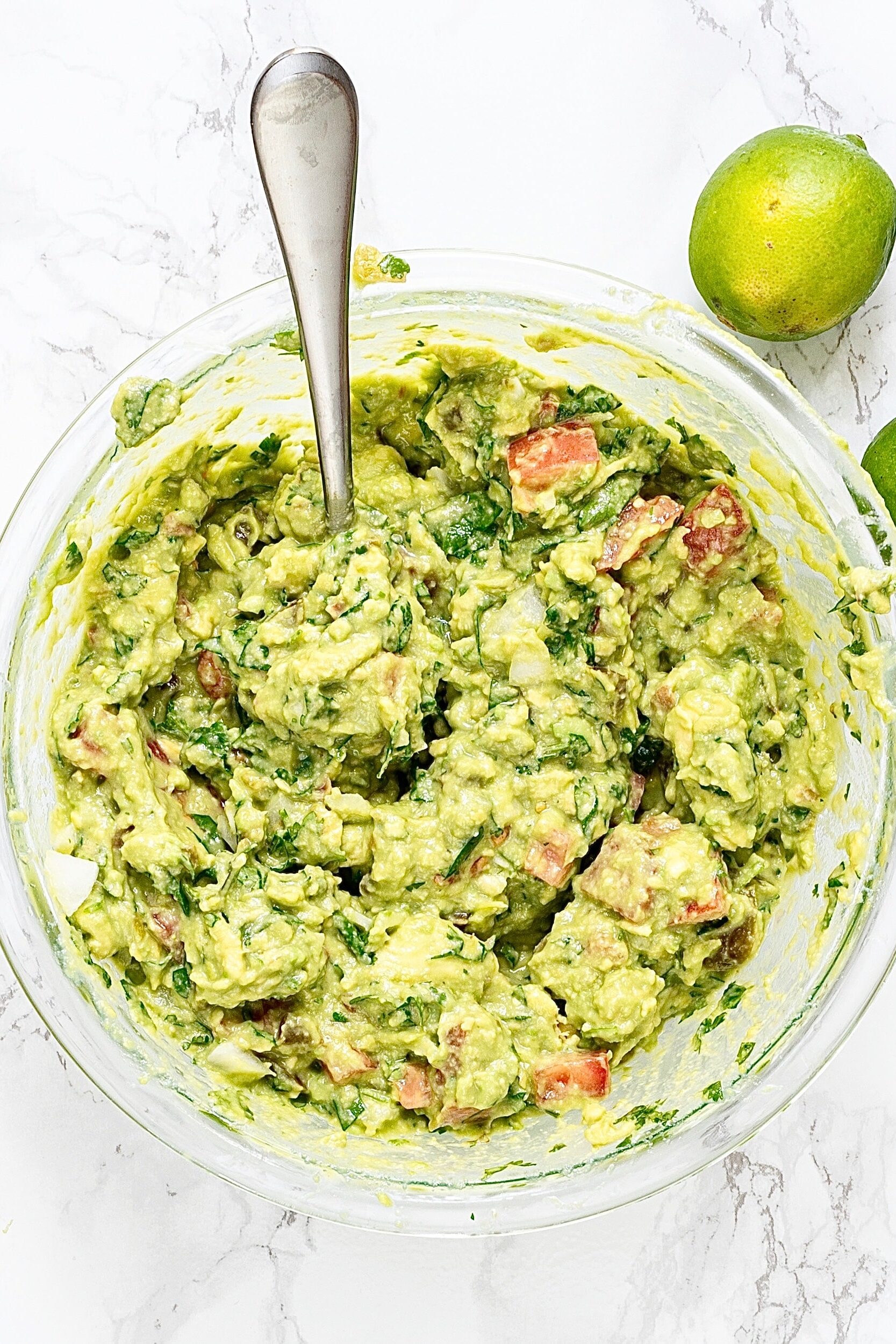 A large glass bowl of guacamole with a serving spoon in it and 2 limes on the counter.