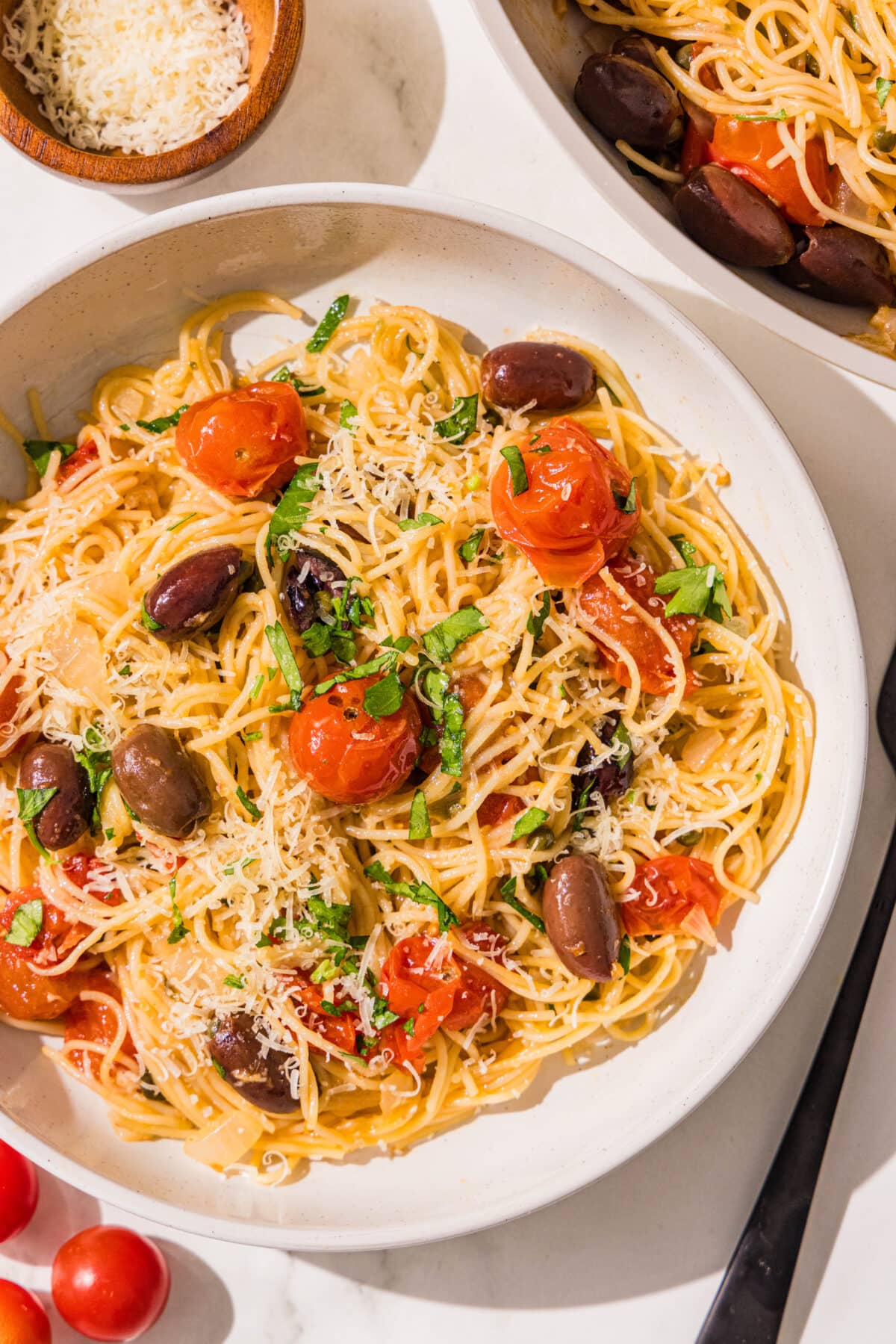 A bowl of spaghetti with tomatoes, kalamata olives, topped with fresh parsley and parmesan. Beside the bowl is a black fork, a couple fresh cherry tomatoes, and a small bowl of parmesan.