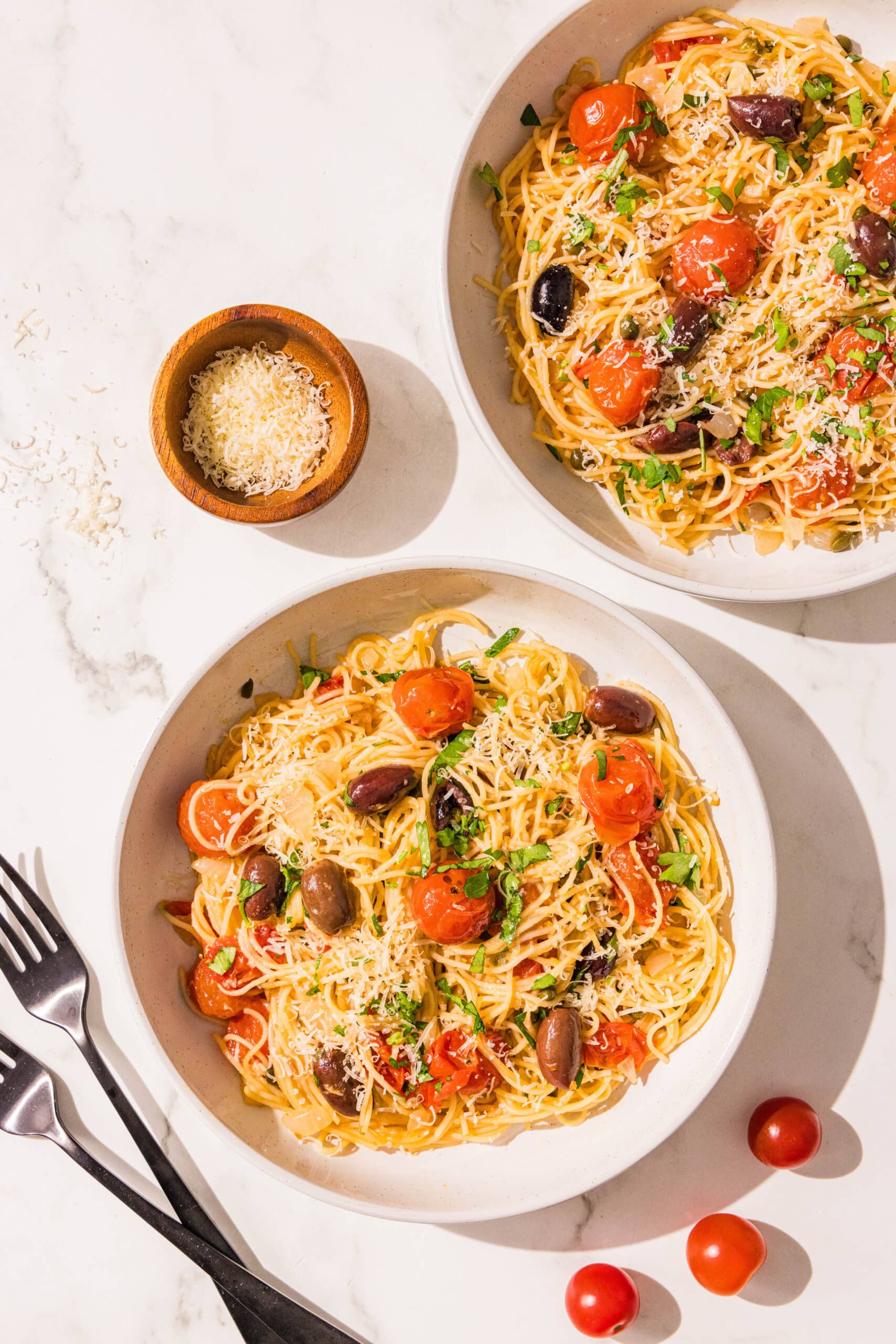 Two bowls of spaghetti with tomatoes and kalamata olives. Beside the bowls are two black forks, a small wooden bowl of grated parmesan, and 3 cherry tomatoes.