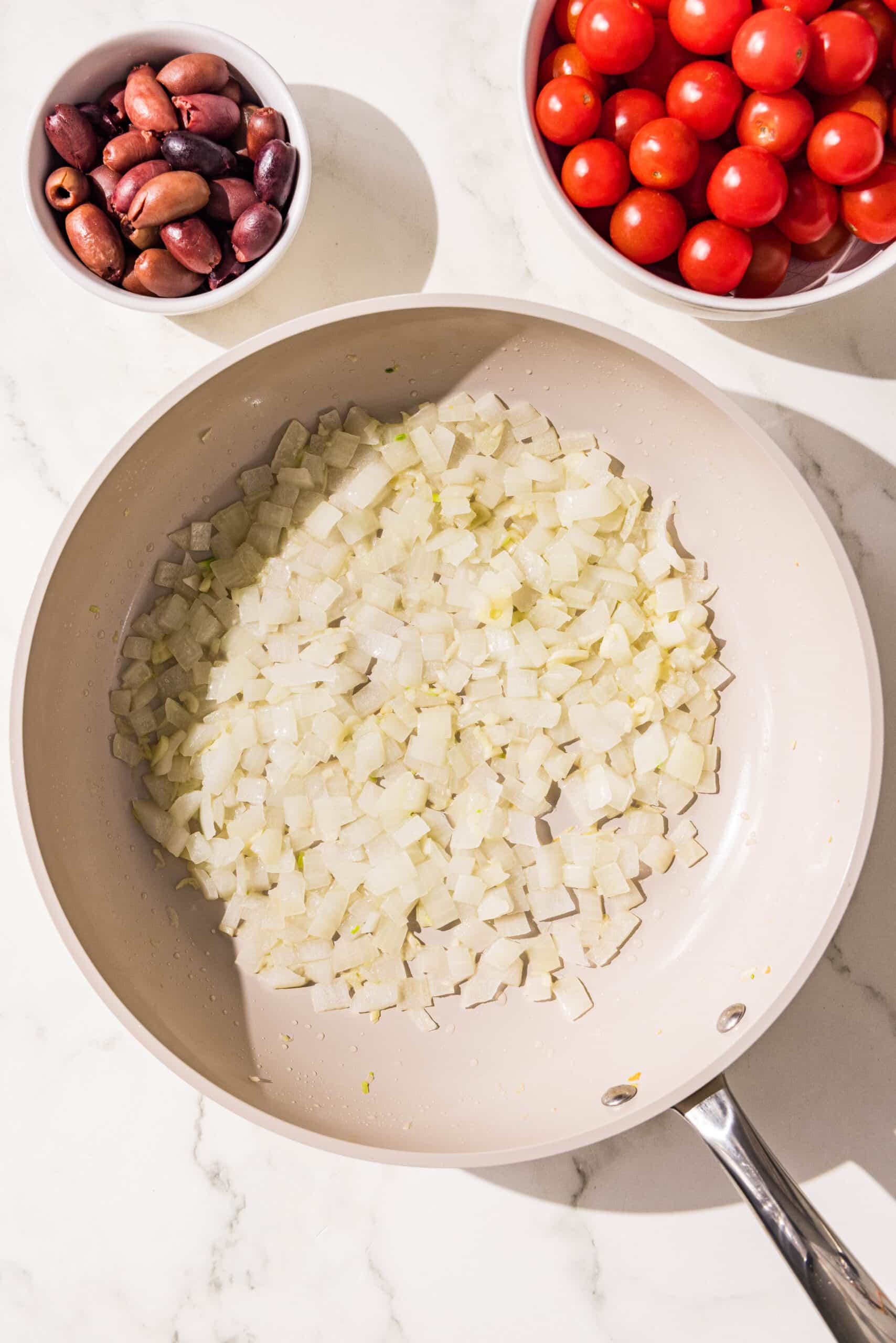 A small pan filled with onions and olive oil along with two small bowls of tomatoes and kalamata olives.