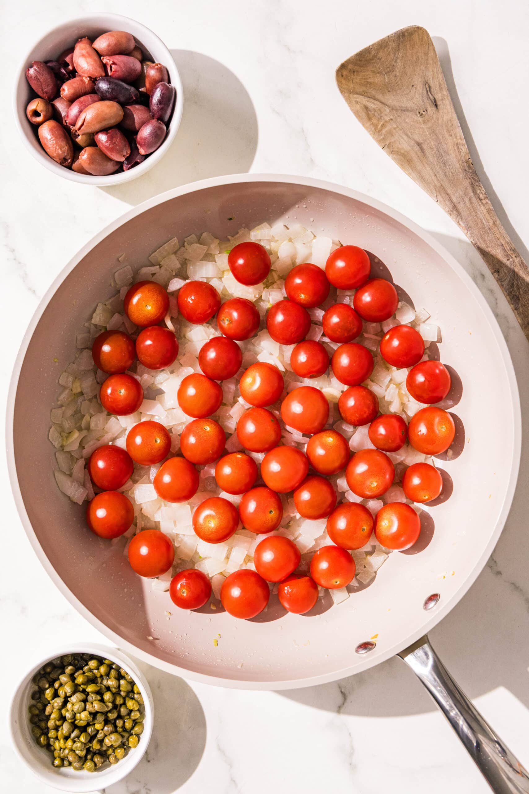 A small pan filled with olive oil, diced onion, and cherry tomatoes. Beside the pan is two small bowls of capers and olives along with a wooden spoon.