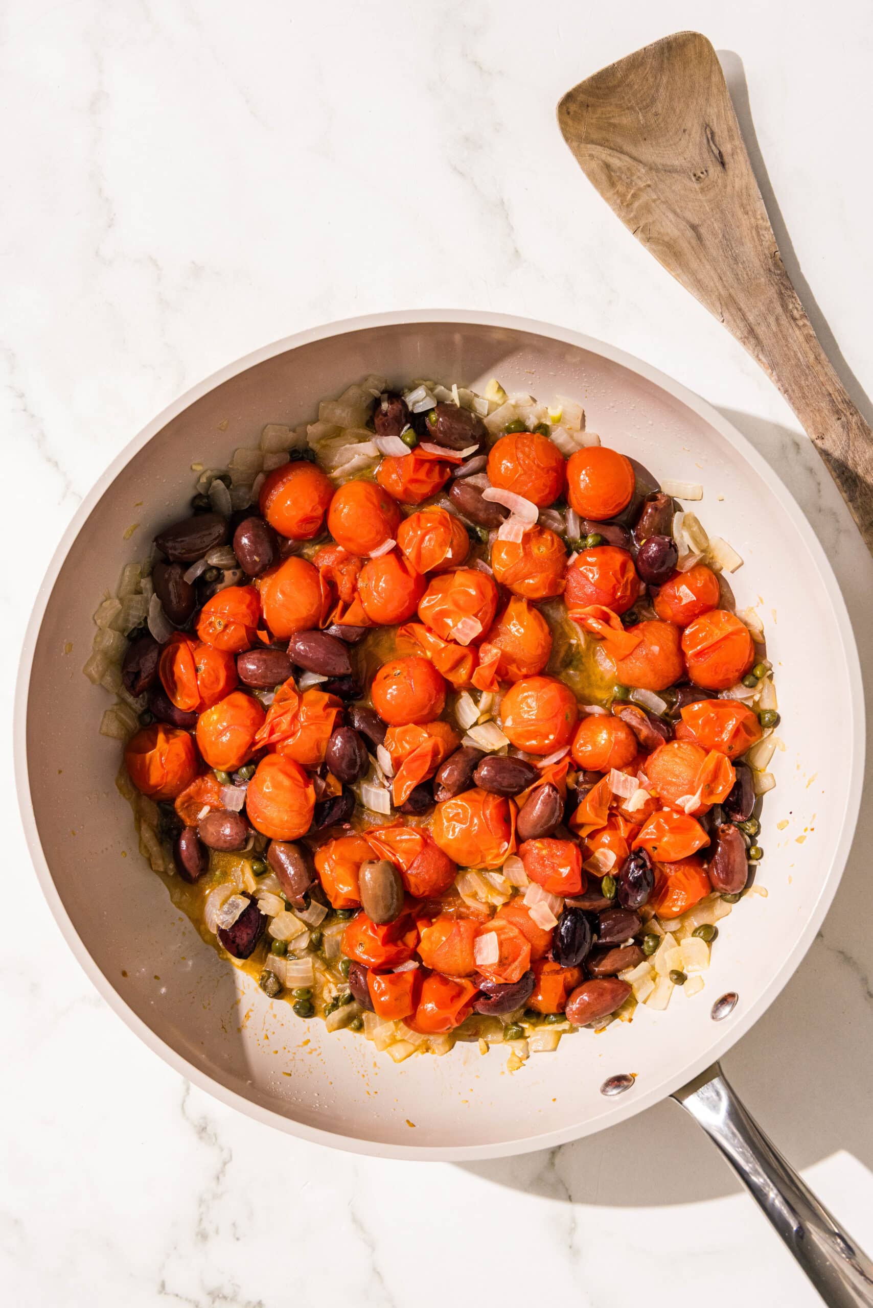 A small pan filled with sauteed onion, kalamata olives, capers, and cherry tomatoes and a wooden spoon.