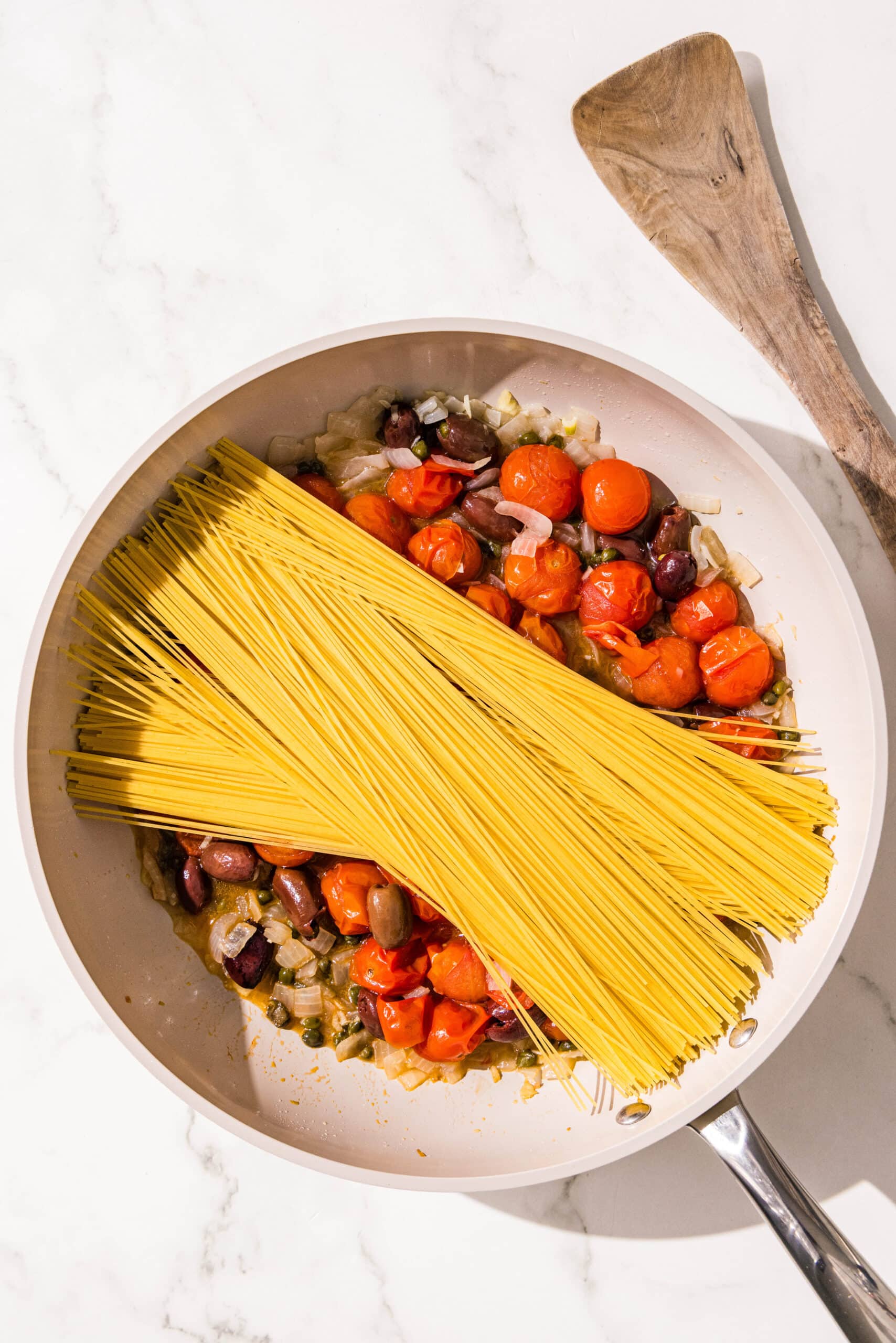 A small pan filled with sauteed onion, kalamata olives, capers, and cherry tomatoes topped with uncooked spaghetti noodles. Beside the pan is a wooden spoon.