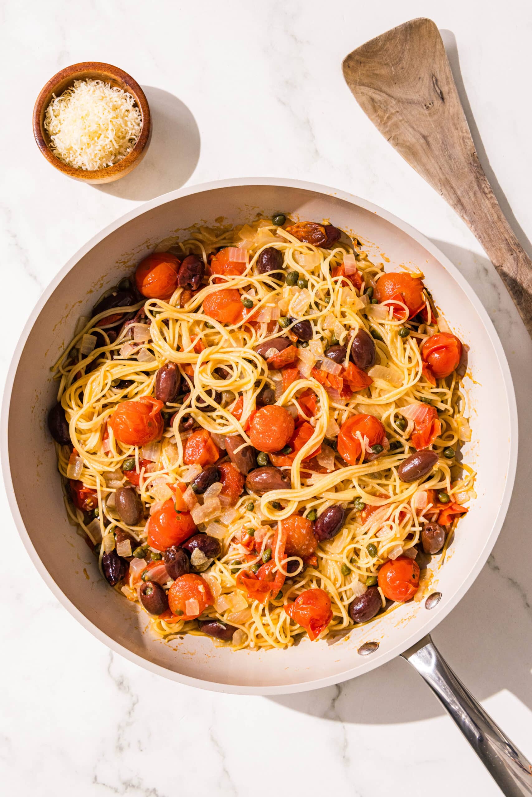 A small pan filled with a spaghetti noodle dish with tomatoes, kalamata olives, and capers. Beside the pan is a small bowl of grated parmesan and a wooden spoon.