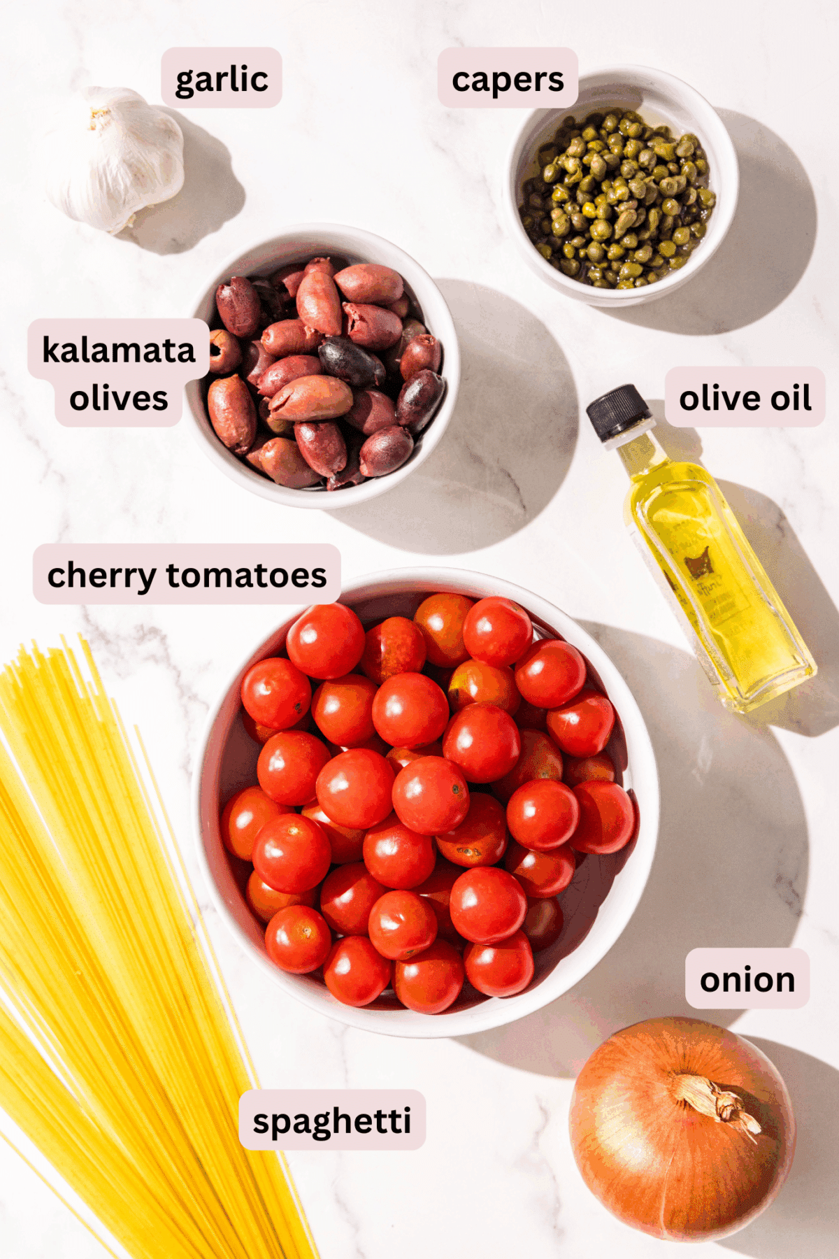 Ingredients laid out on a counter in bowls and labeled including: garlic, capers, kalamata olives, olive oil, cherry tomatoes, spaghetti noodles, and onion.