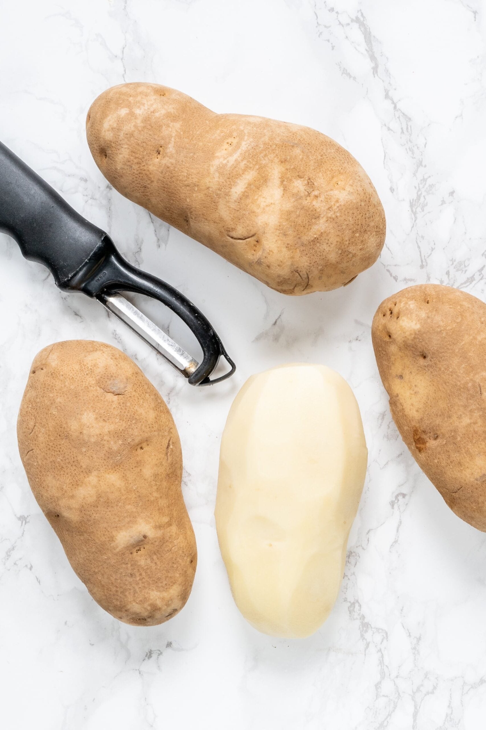 Four russet potatoes, one of them is peeled and a black peeler.