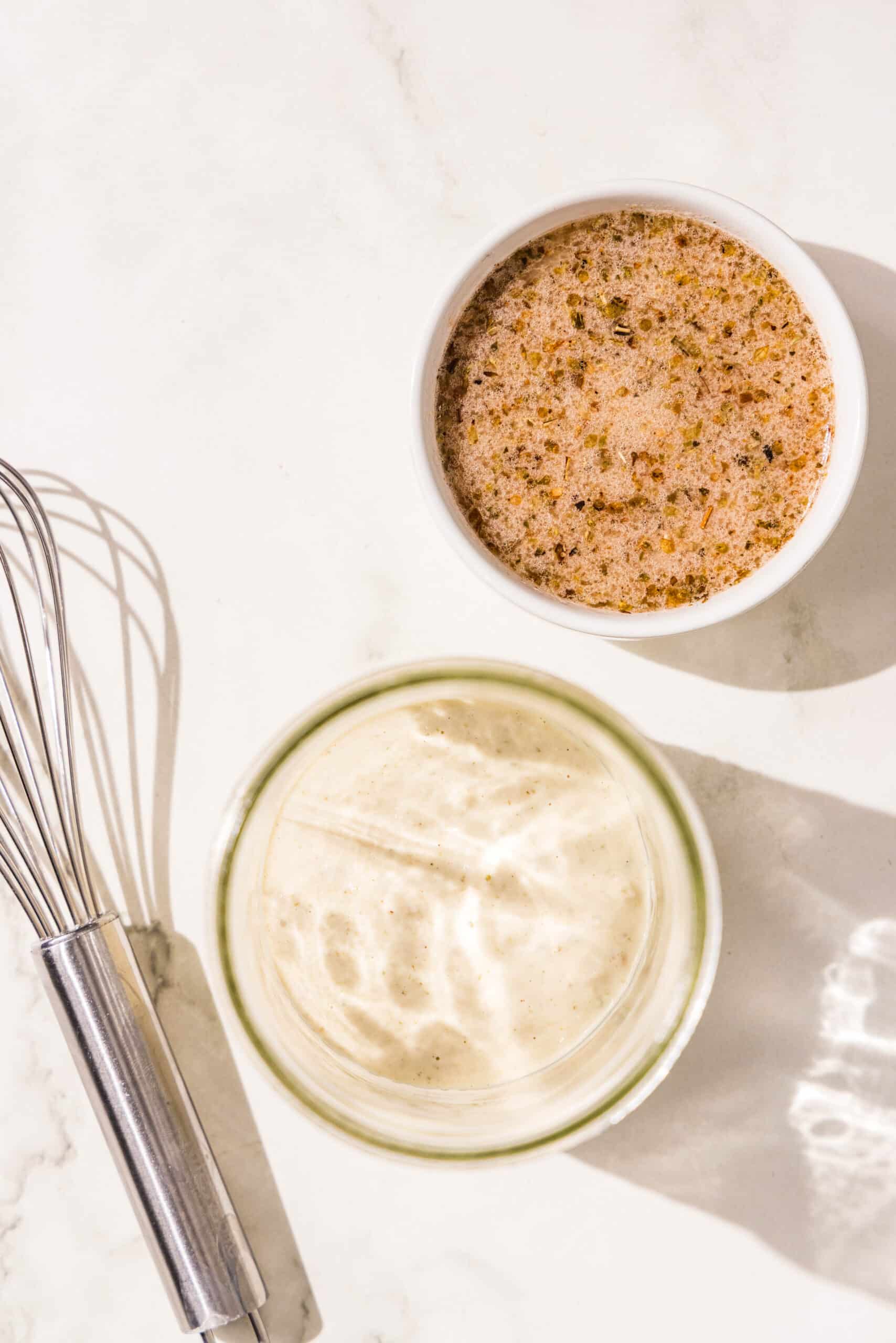 Two bowls of salad dressing and a metal whisk.