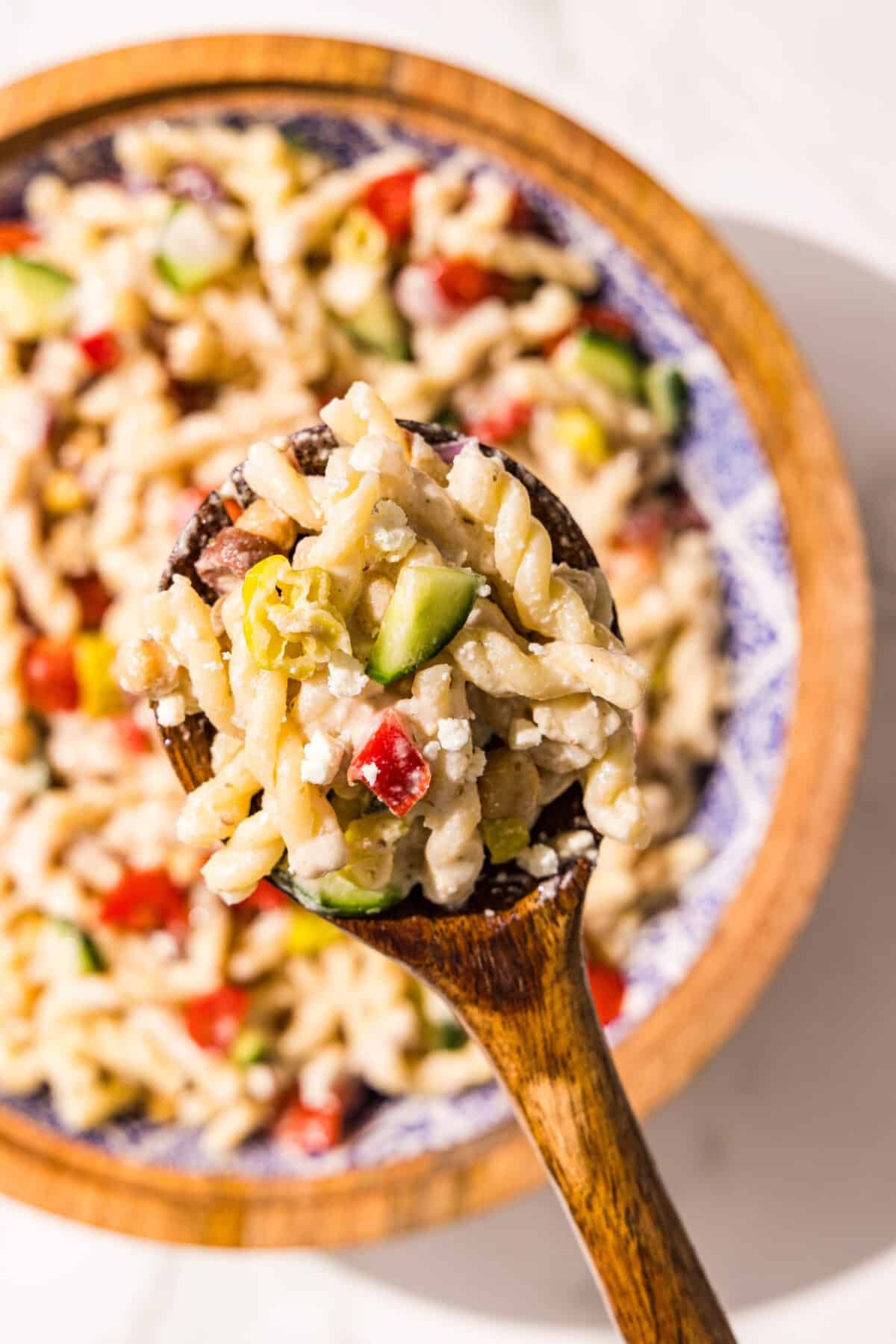 A large bowl with greek pasta salad and a wooden spoon holding a scoop of pasta salad.