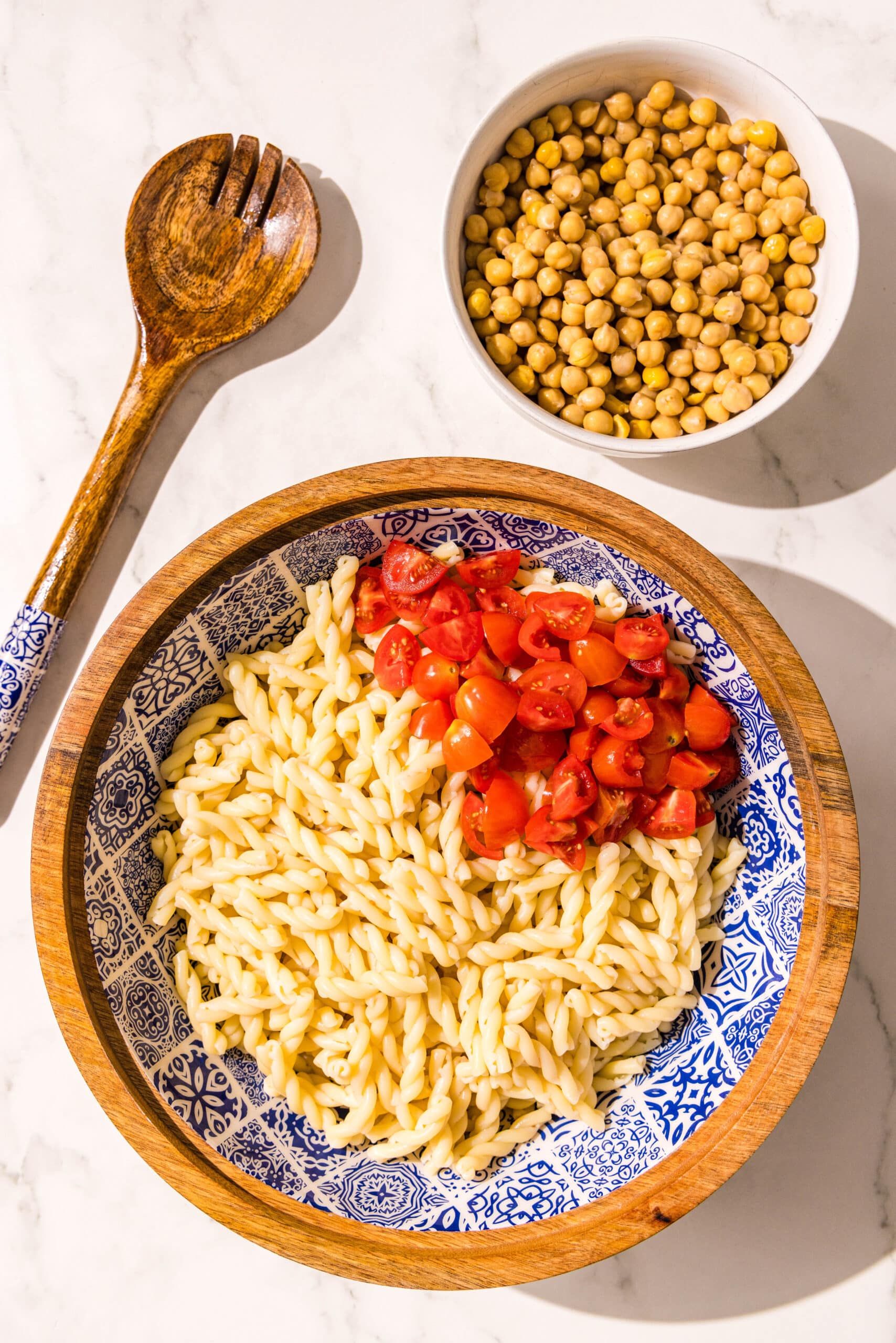 A large wooden bowl filled with pasta noodles and halved tomatoes. Beside the bowl is a smaller bowl of chickpeas and a wooden spoon.