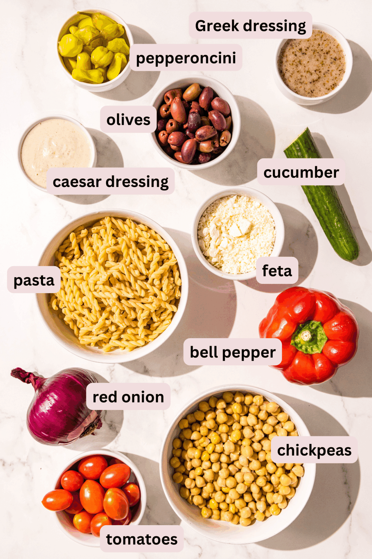 Ingredients laid out on a counter in bowls and labeled including greek dressing, caesar dressing, pepperonicini, cucumber, feta, pasta, bell pepper, red onion, chickpeas, and tomatoes.