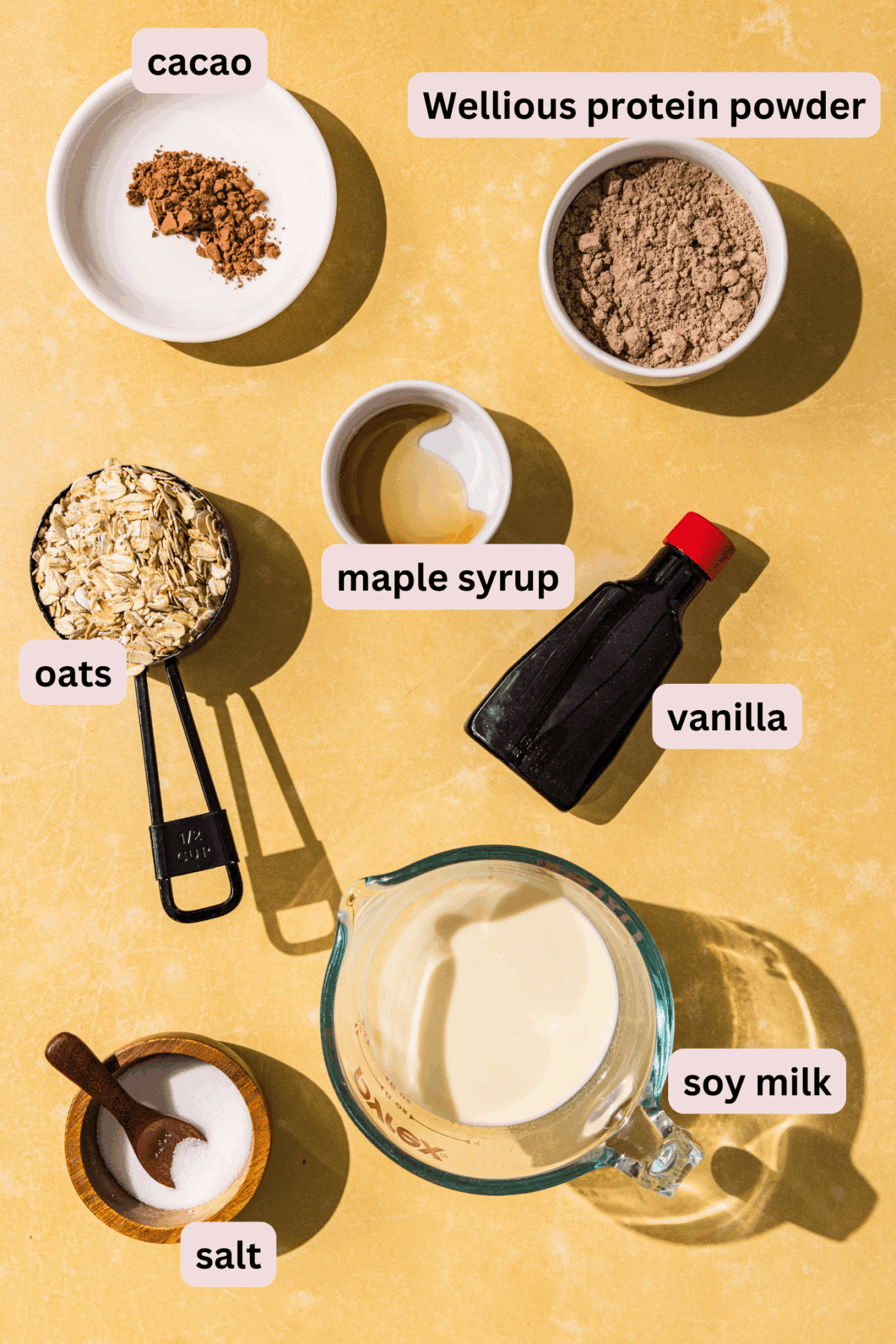 Ingredients laid out on a yellow countertop and labeled including: cacao, protein powder, maple syrup, vanilla, oats, soy milk, and salt.