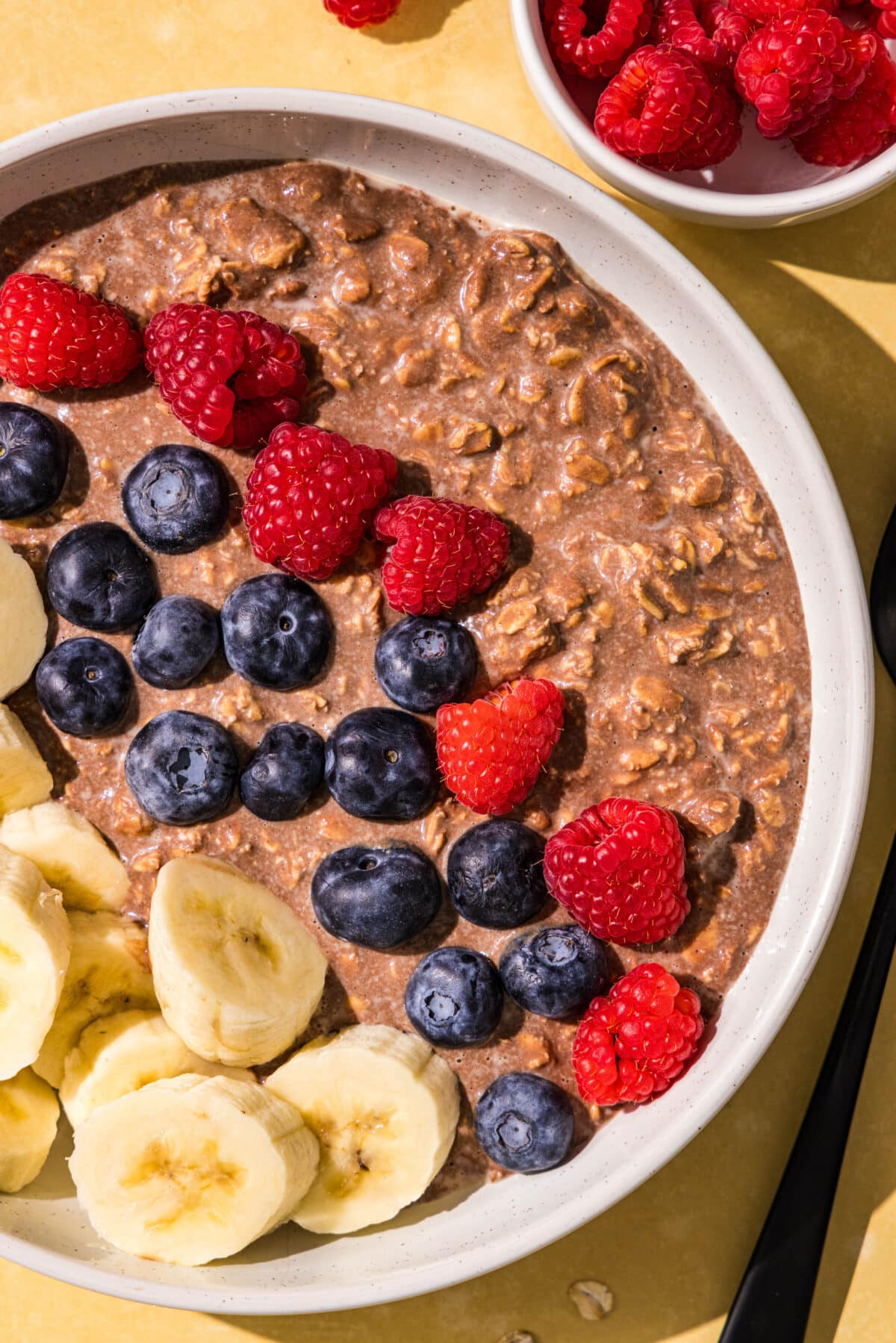 A bowl of chocolate oats topped with raspberries, blueberries, and sliced banana. Beside the bowl is a black spoon and a small bowl of raspberries.