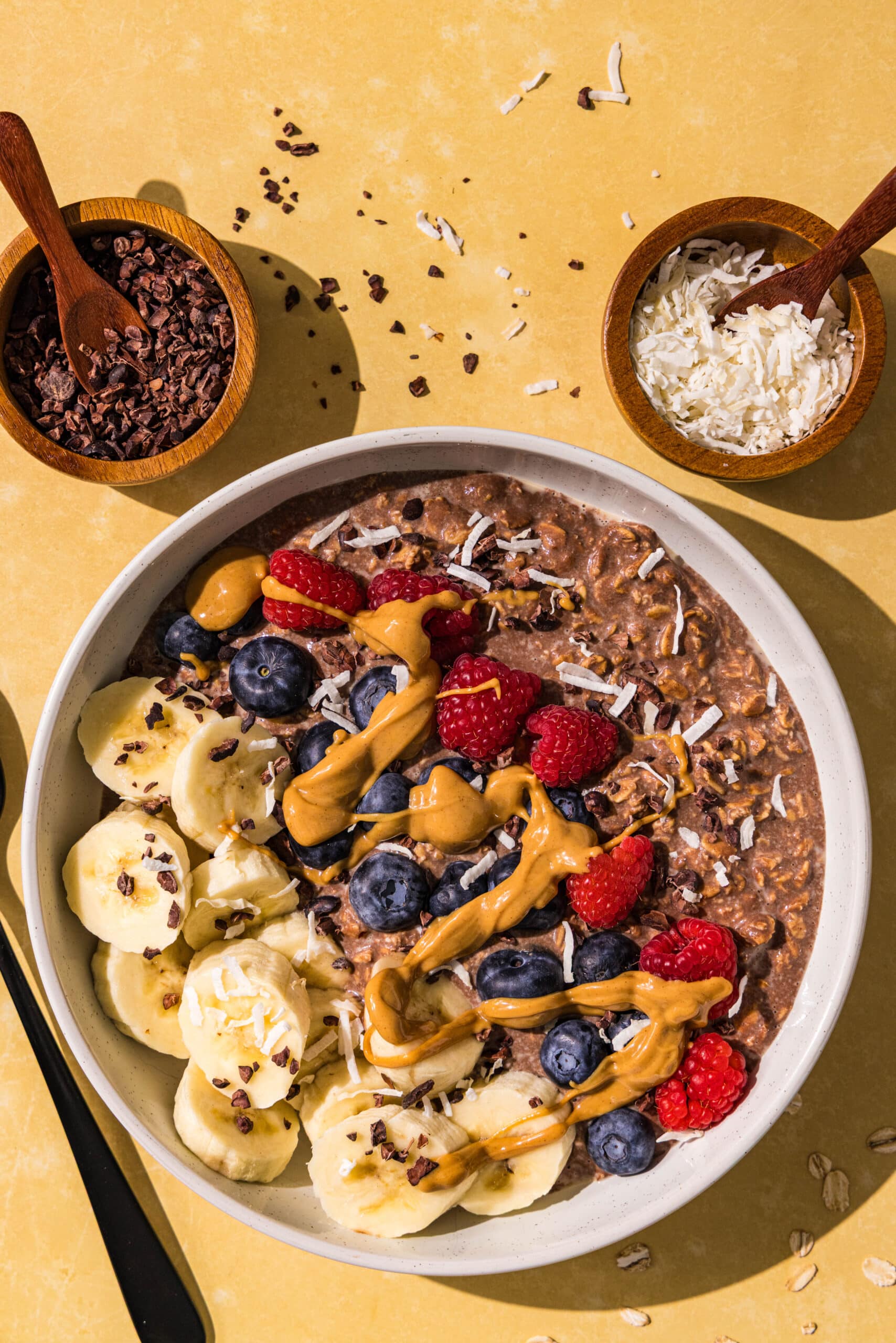 A bowl of chocolate oatmeal topped with raspberries, blueberries, sliced banana, drizzle with peanut butter and sprinkled with cacao nibs and shredded coconut. Beside the bowl is two small bowls of raspberries and cacao nibs.
