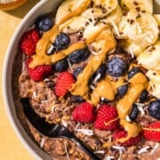A bowl of chocolate oatmeal topped with raspberries, blueberries, sliced banana, drizzle with peanut butter and sprinkled with cacao nibs and shredded coconut. Beside the bowl is two small bowls of raspberries and coconut.