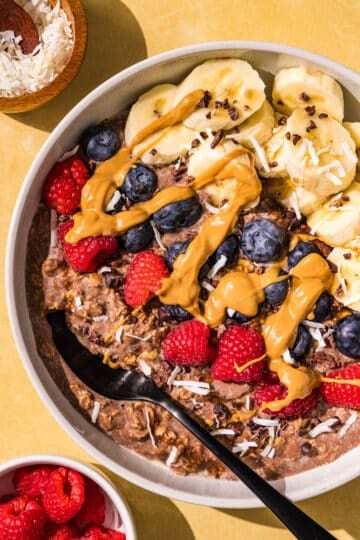A bowl of chocolate oatmeal topped with raspberries, blueberries, sliced banana, drizzle with peanut butter and sprinkled with cacao nibs and shredded coconut. Beside the bowl is two small bowls of raspberries and coconut.