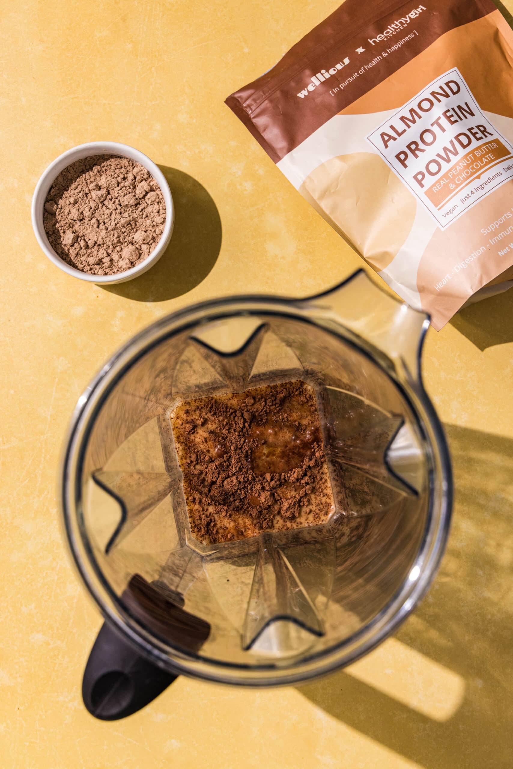 A blender filled with soy milk and cacao powder, a bag of protein powder, and a small bowl of protein powder.