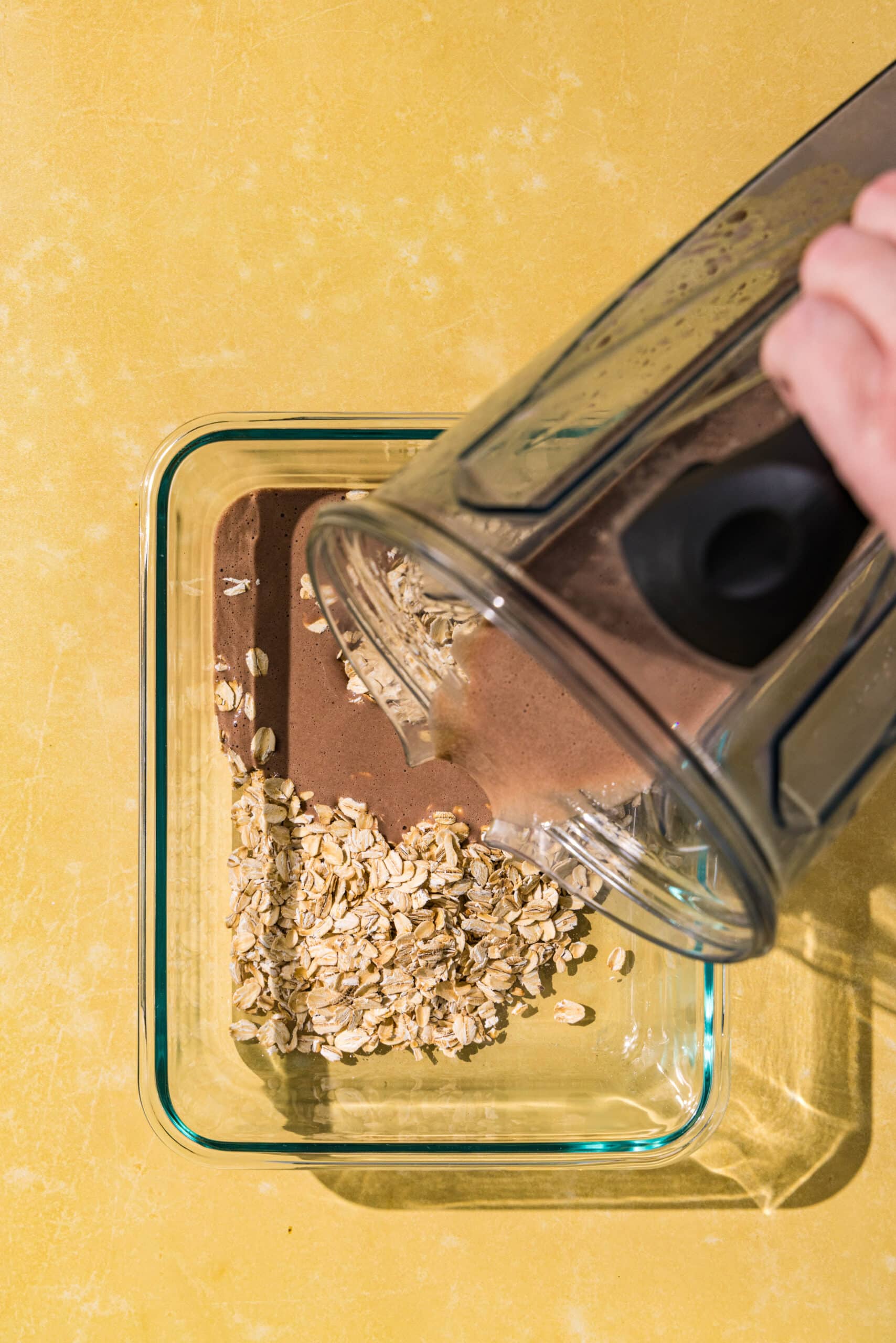 A glass dish filled with oats and a blender pouring chocolate milk over the oats.