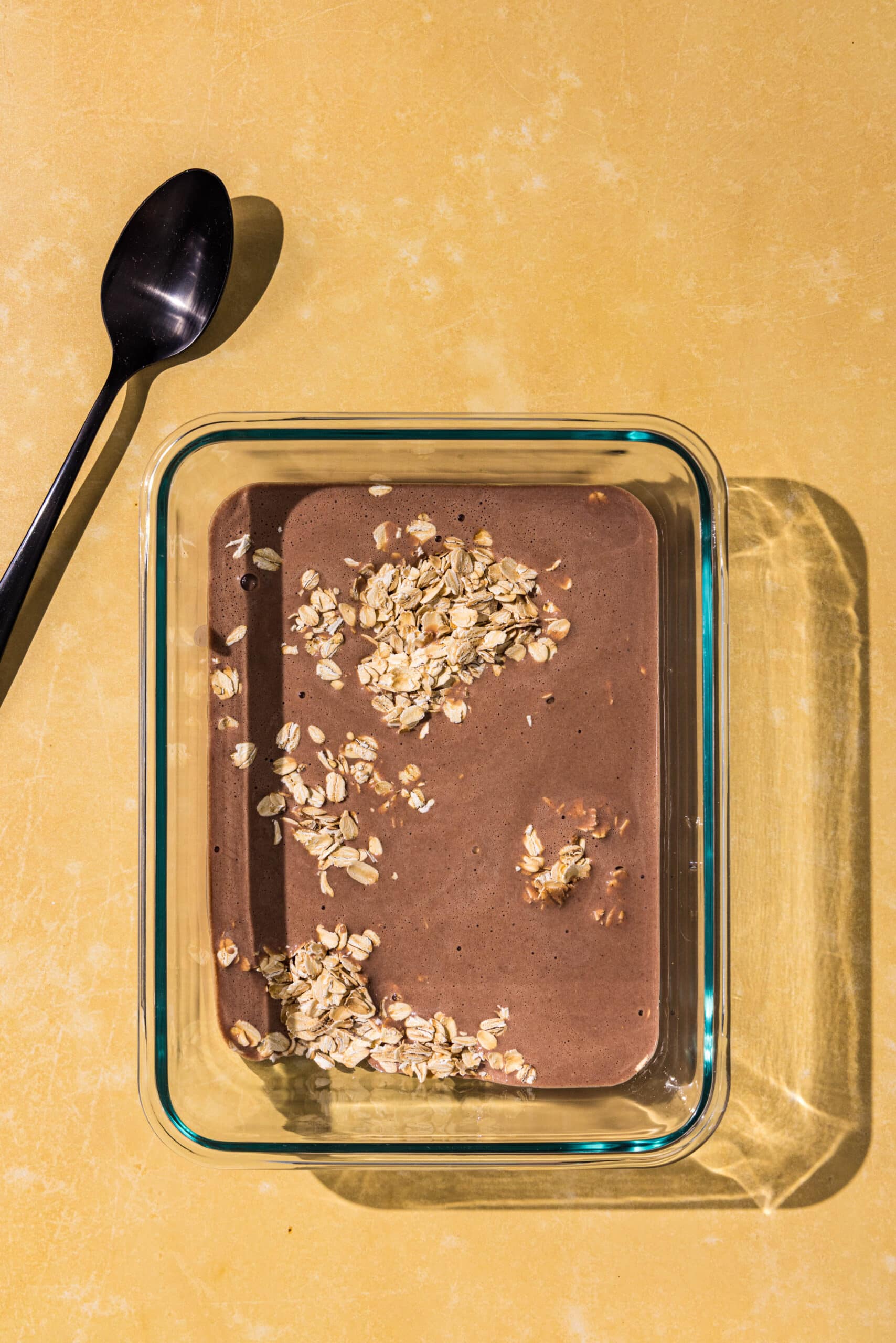 A glass dish filled with oats and chocolate milk and a black spoon.