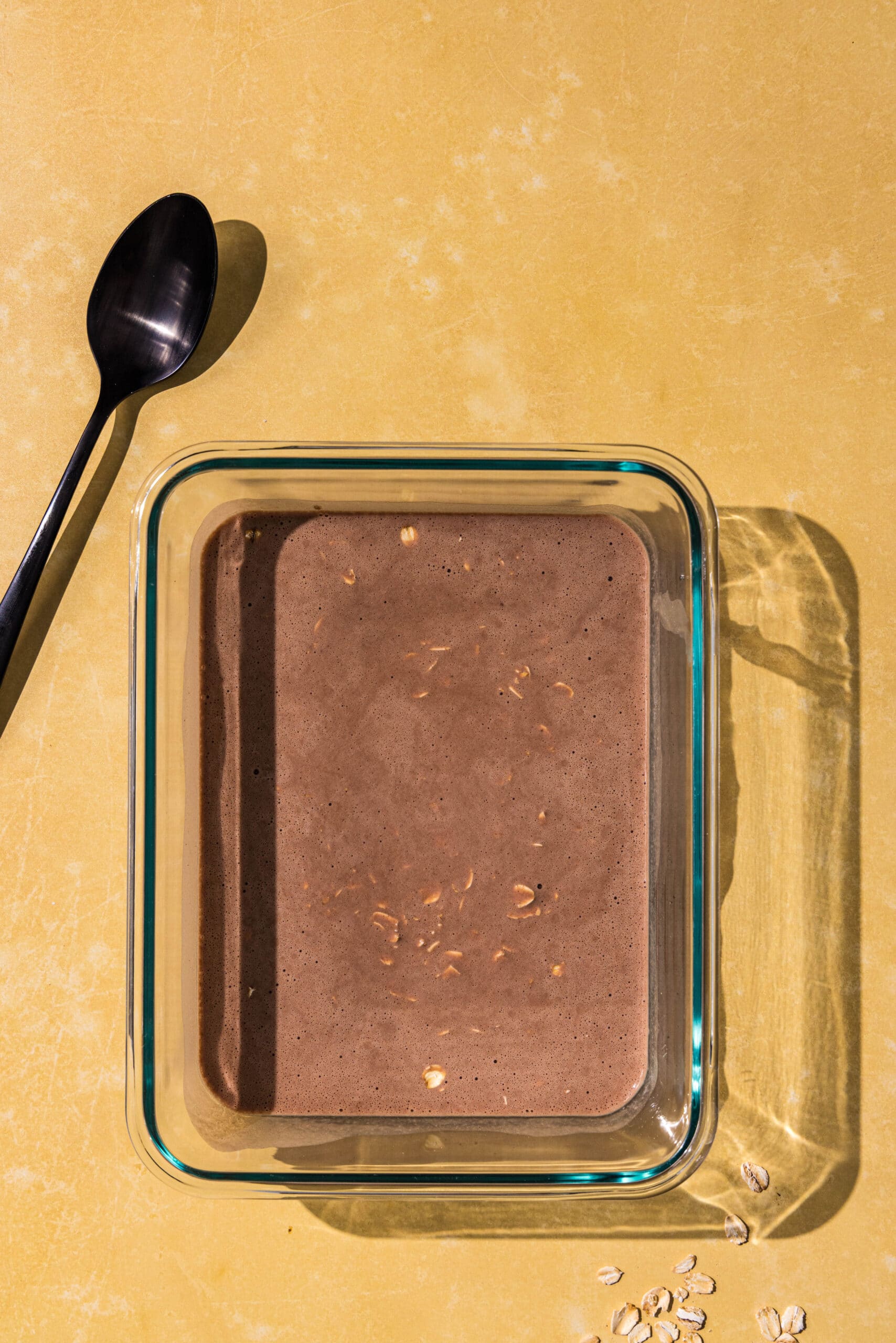 A glass dish filled with chocolate milk and oats and a black spoon.