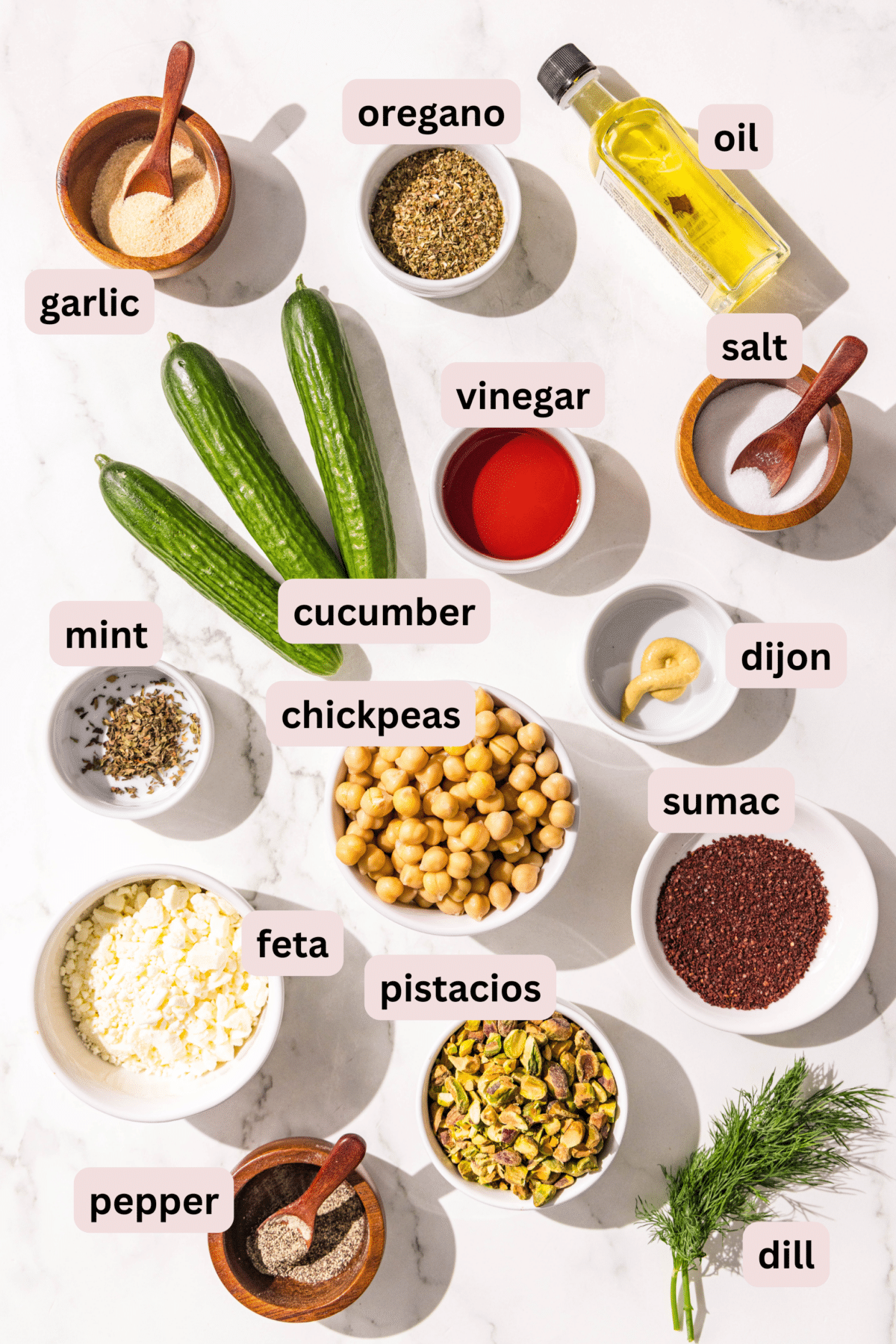 Ingredients laid out on a marble countertop and labeled including: garlic powder, oregano, oil, cucumbers, vinegar, salt, mint, chickpeas, Dijon mustard, feta, pistachios, sumac, pepper, and dill.