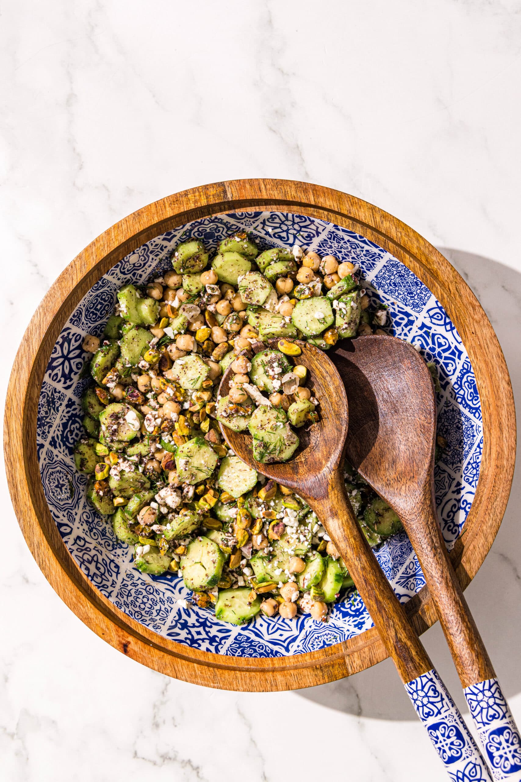 A large bowl of cucumber feta chickpea salad and a pair of wooden tongs.