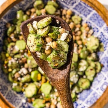 A big bowl of cucumber chickpea salad and a wooden spoon holding a scoop of the salad.