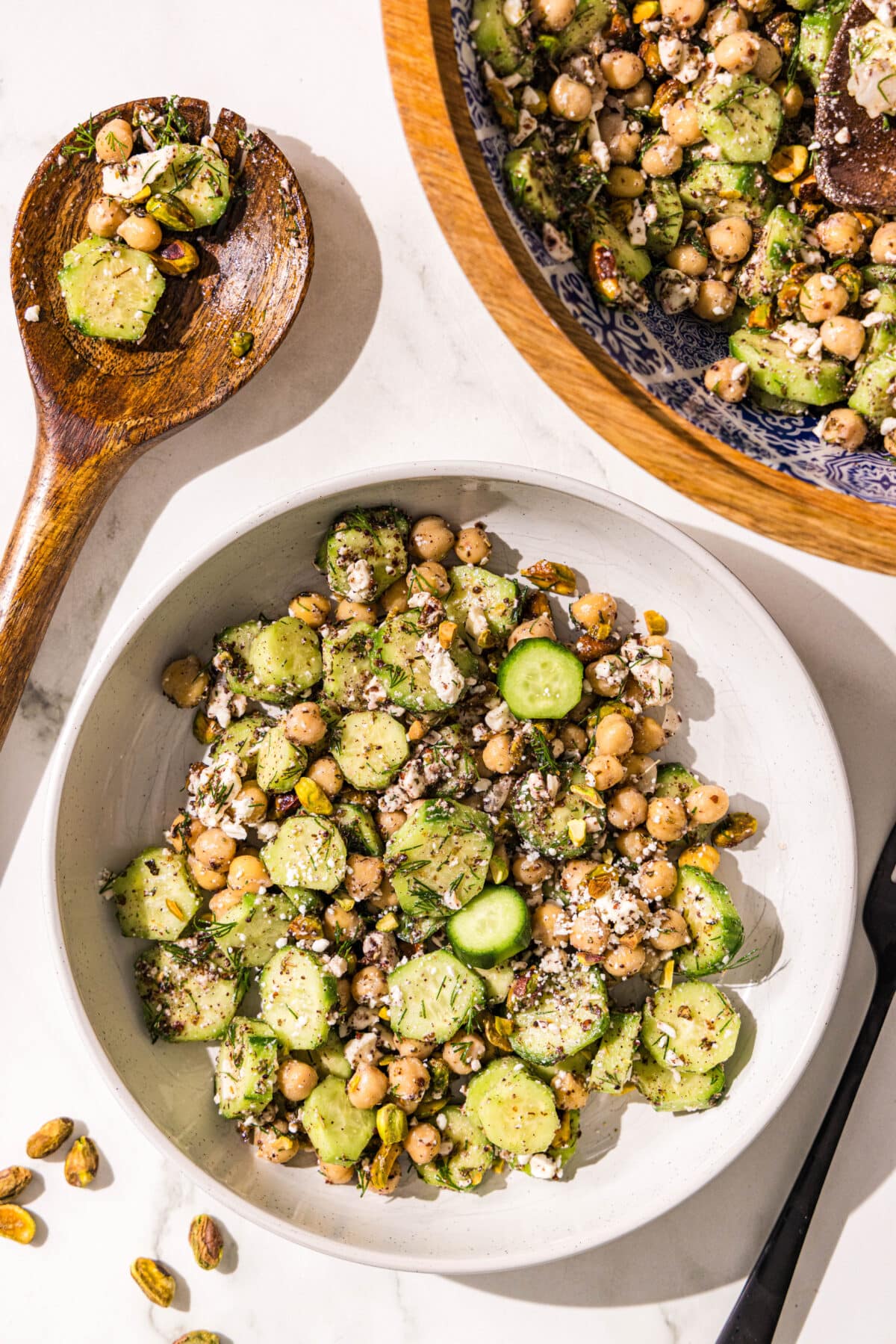 A white bowl filled with cucumber chickpea feta salad, a wooden spoon, a black fork, and a wooden bowl with remaining salad.