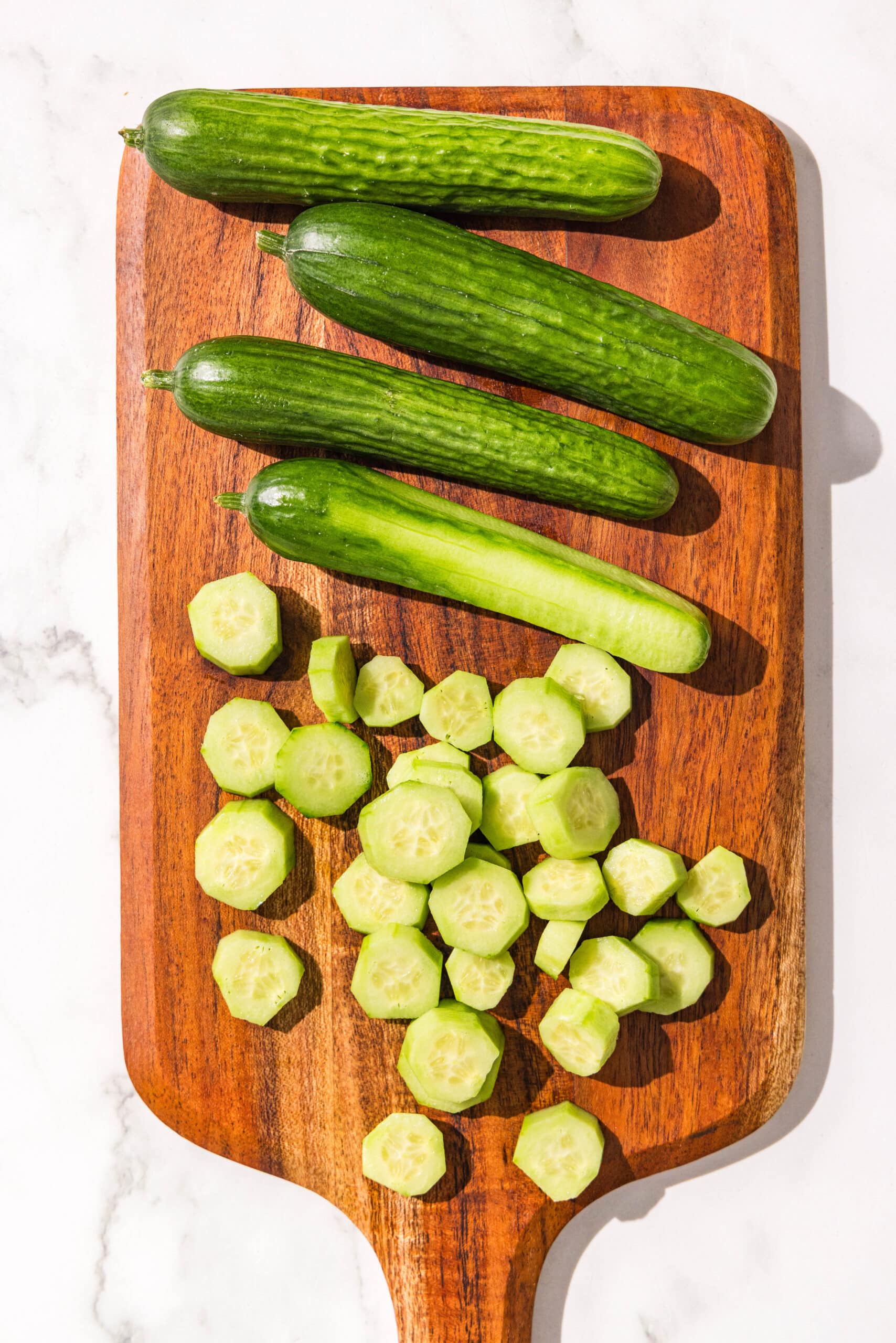 A wooden cutting board with 4 whole cucumbers and sliced cucumbers on it.