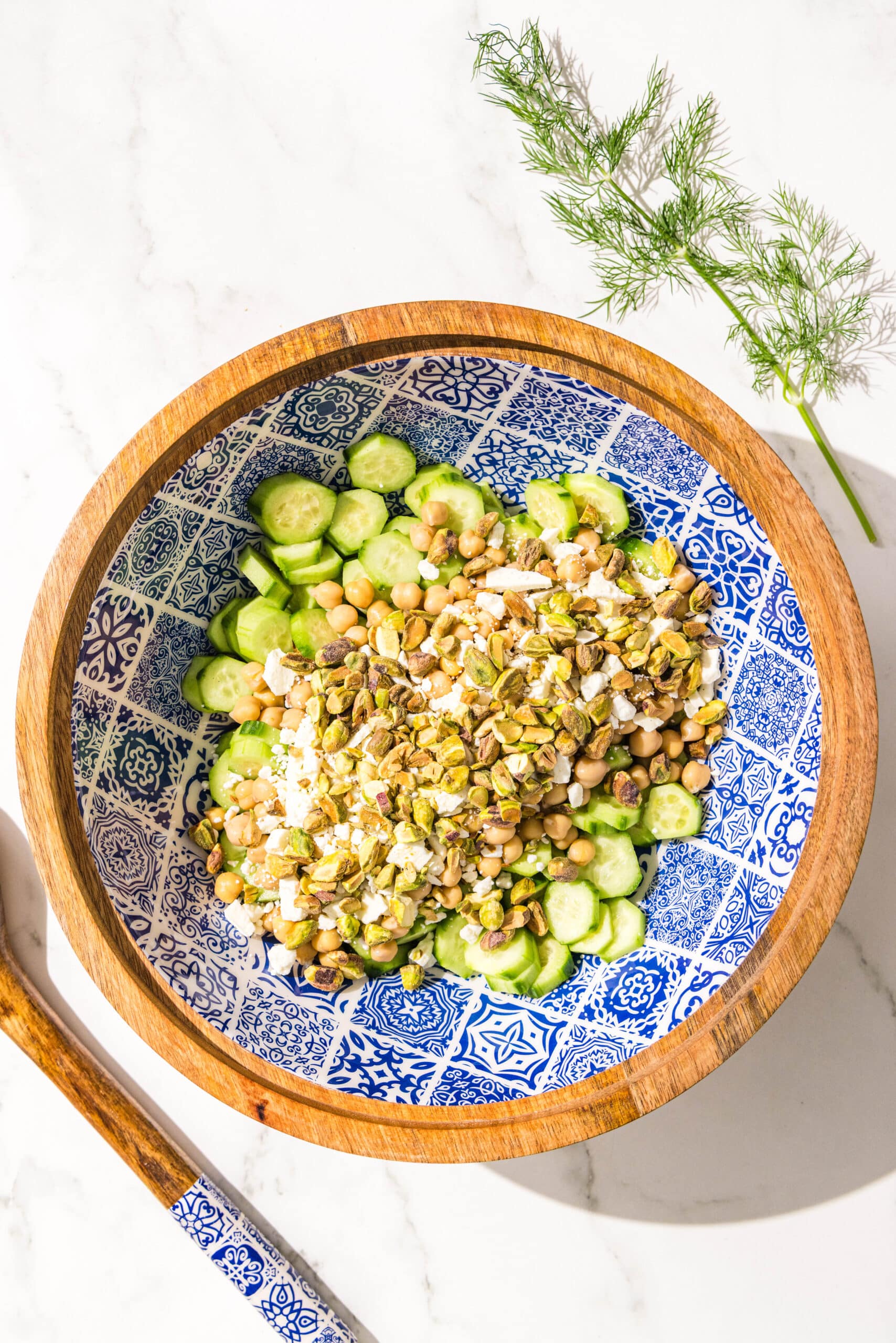 A large bowl filled with cucumbers, chickpeas, feta, and pistachios. Plus a sprig of fresh dill next to the bowl.