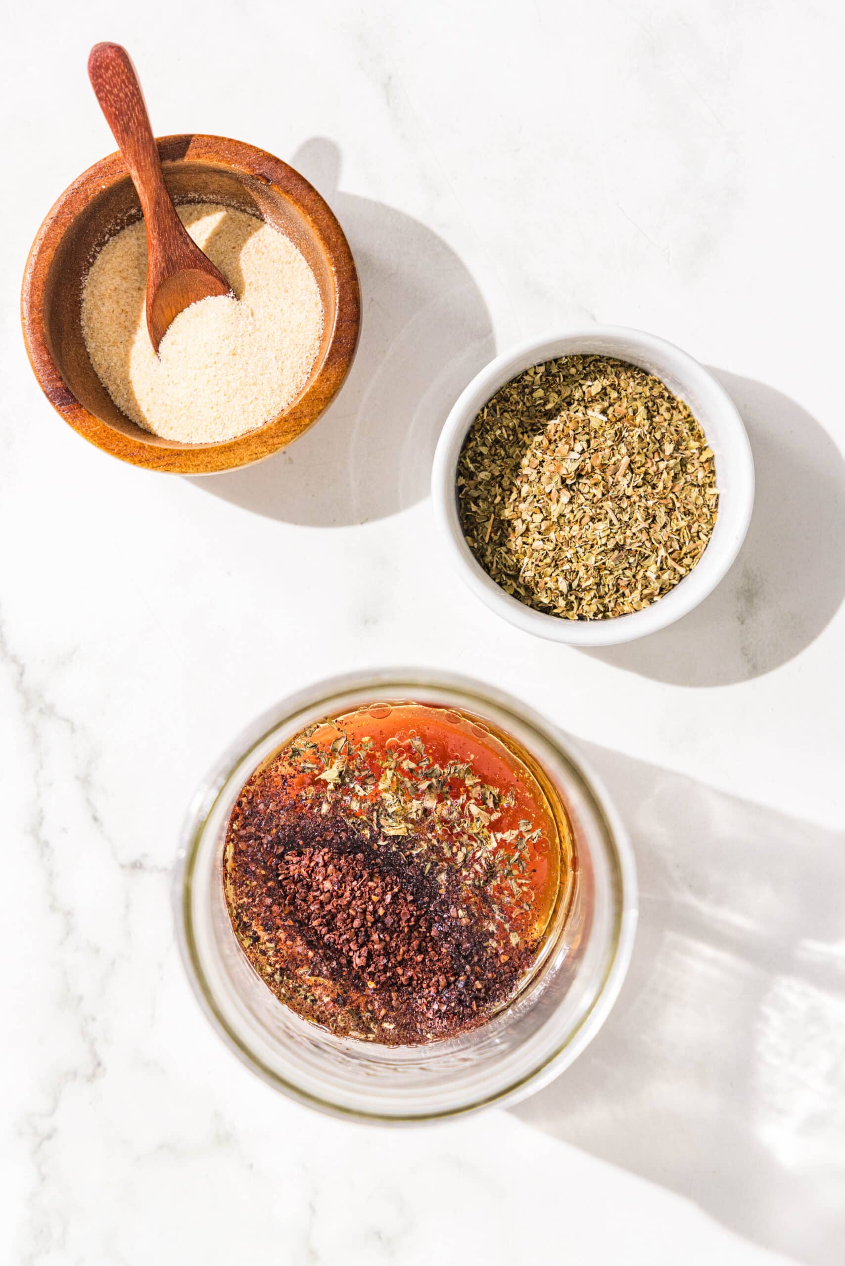 A glass jar filled with olive oil, red wine vinegar, and spices, plus two bowls with garlic powder and oregano in them.