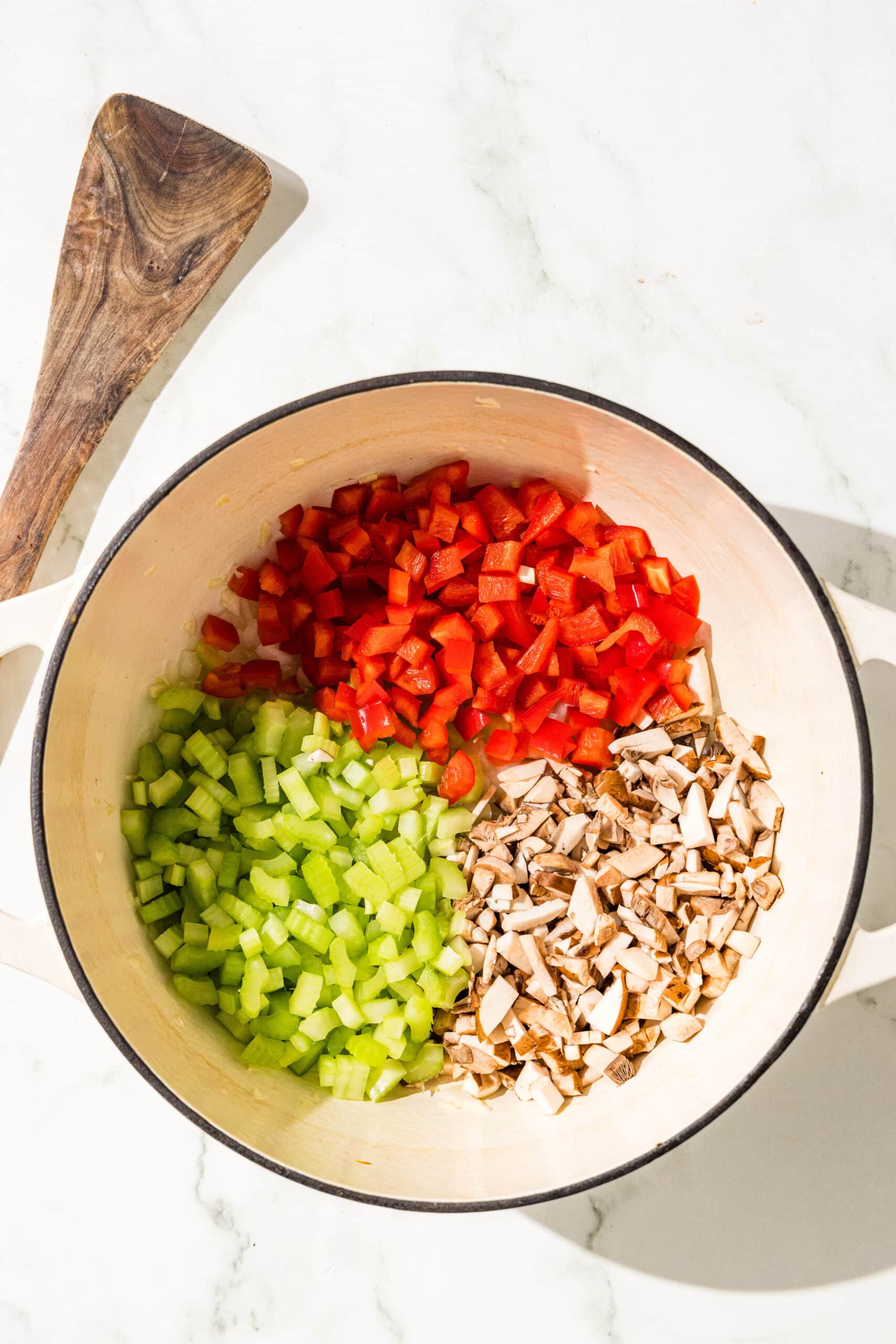 A soup pot with chopped mushrooms, bell pepper, and celery in it and a wooden spoon.