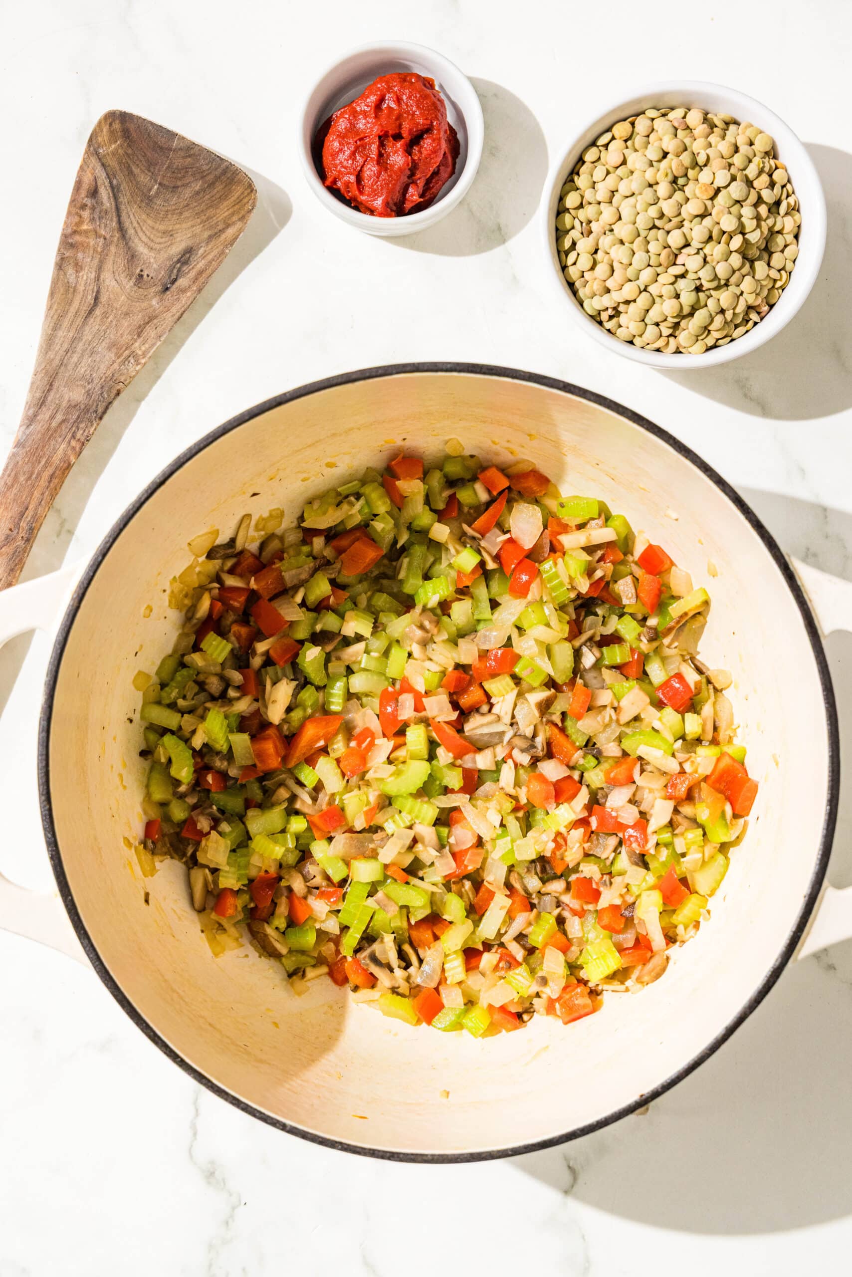 A soup pot with sauteed peppers, onion, mushrooms, celery and garlic. Beside the pot is a wooden spoon and two bowls of dried green lentils and tomato paste.