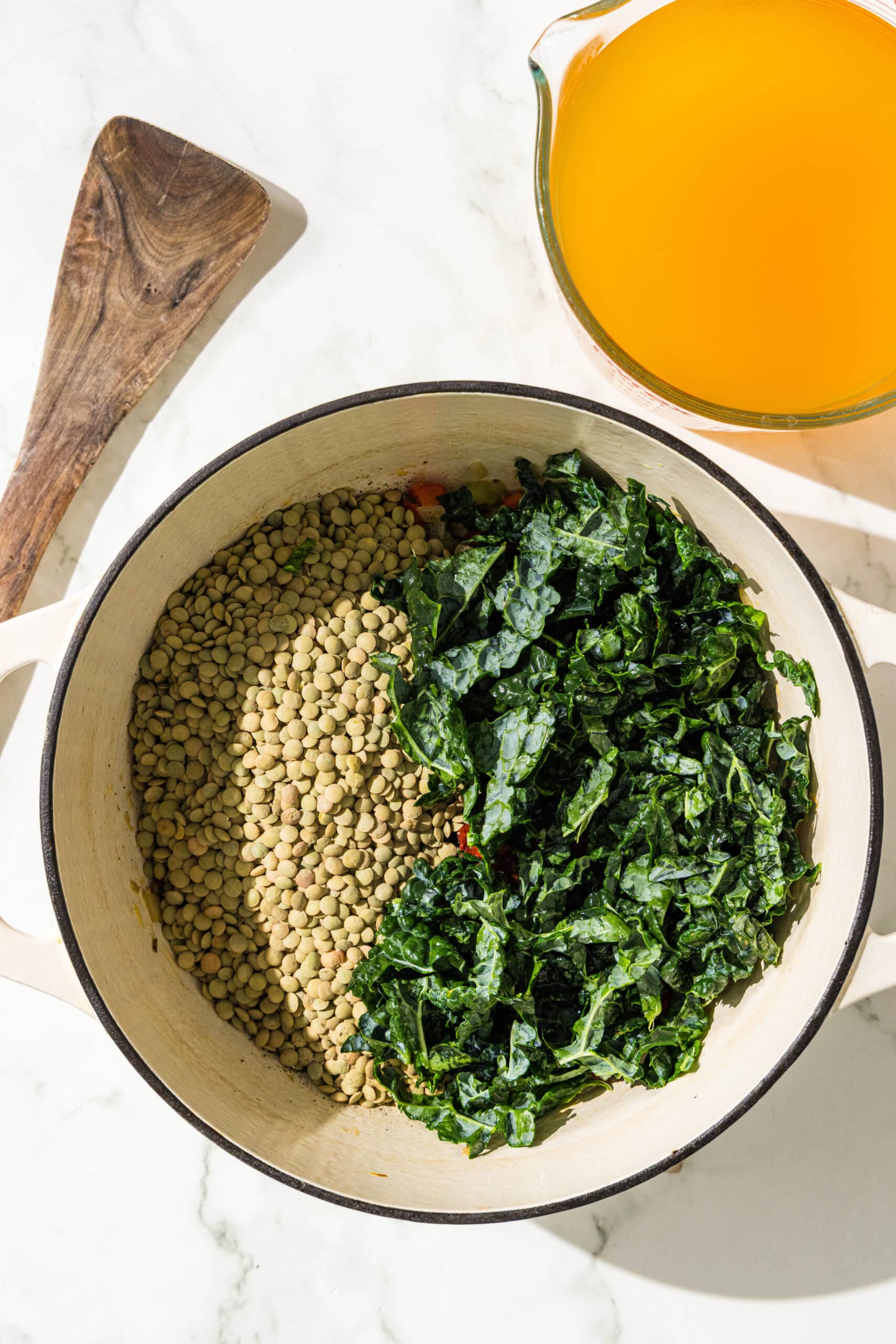 A soup pot with lentils and kale in it, a wooden spoon, and a pitcher of vegetable broth.