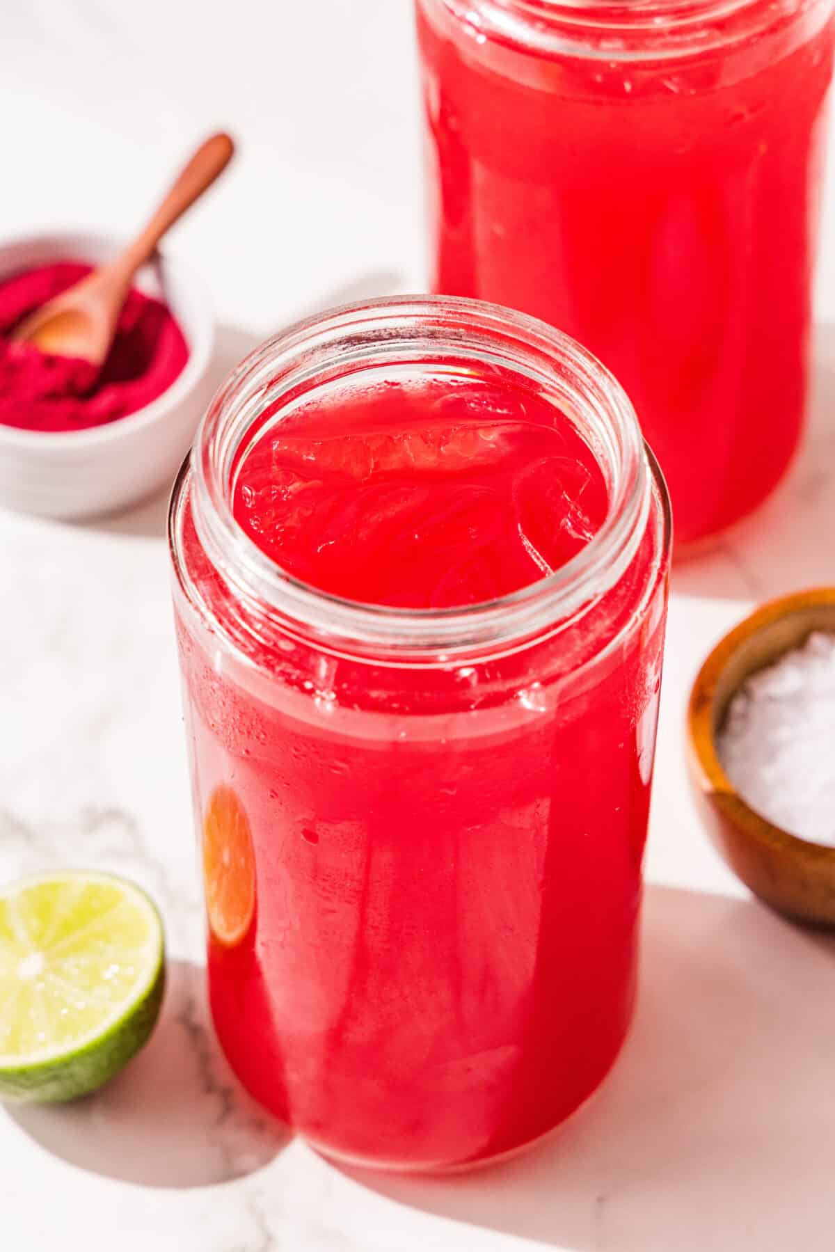 Two glasses of red juice with ice, half of a lime, and two bowls of salt and beet powder.