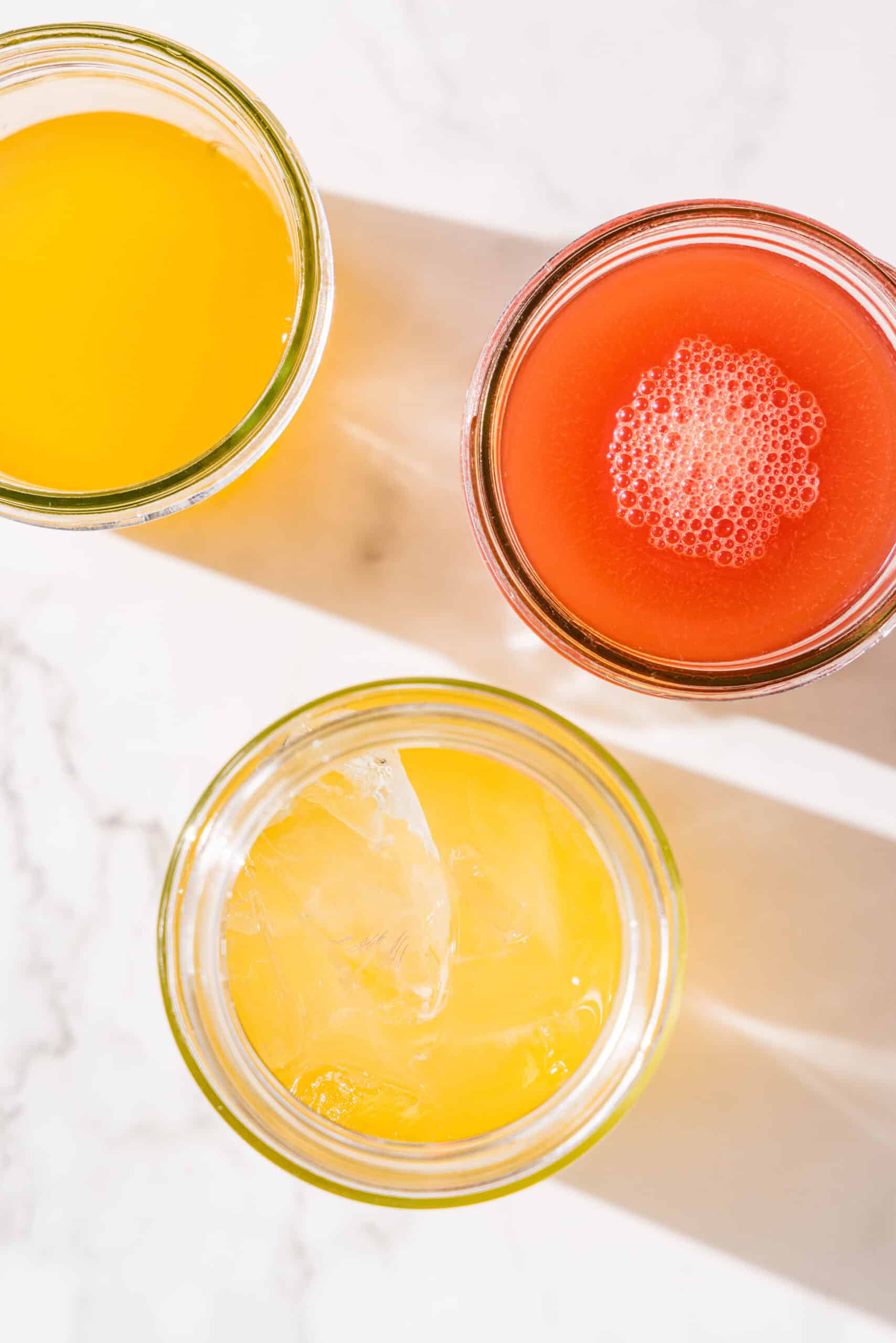 Three glass jars one filled with orange juice, one with watermelon juice, and one with orange juice and ice cubes.