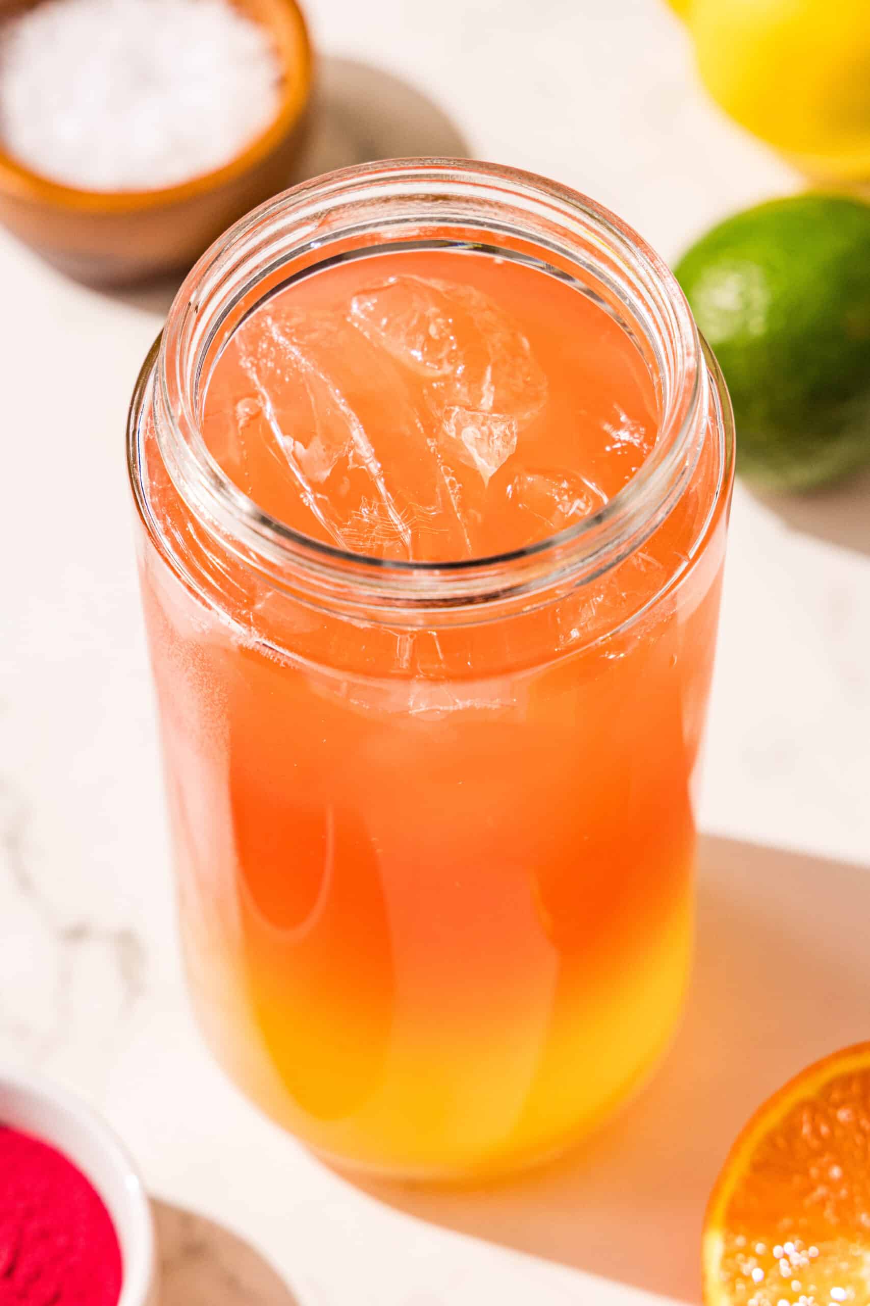 A glass jar filled with ice, orange juice and watermelon juice. Behind the jar is a lemon, a lime, and a bowl of salt.