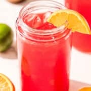 A glass of red juice filled with ice and garnished with an orange slice. On the counter around the glass is a lime, a lemon, half of an orange, and a bowl of salt.