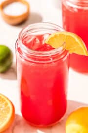A glass of red juice filled with ice and garnished with an orange slice. On the counter around the glass is a lime, a lemon, half of an orange, and a bowl of salt.