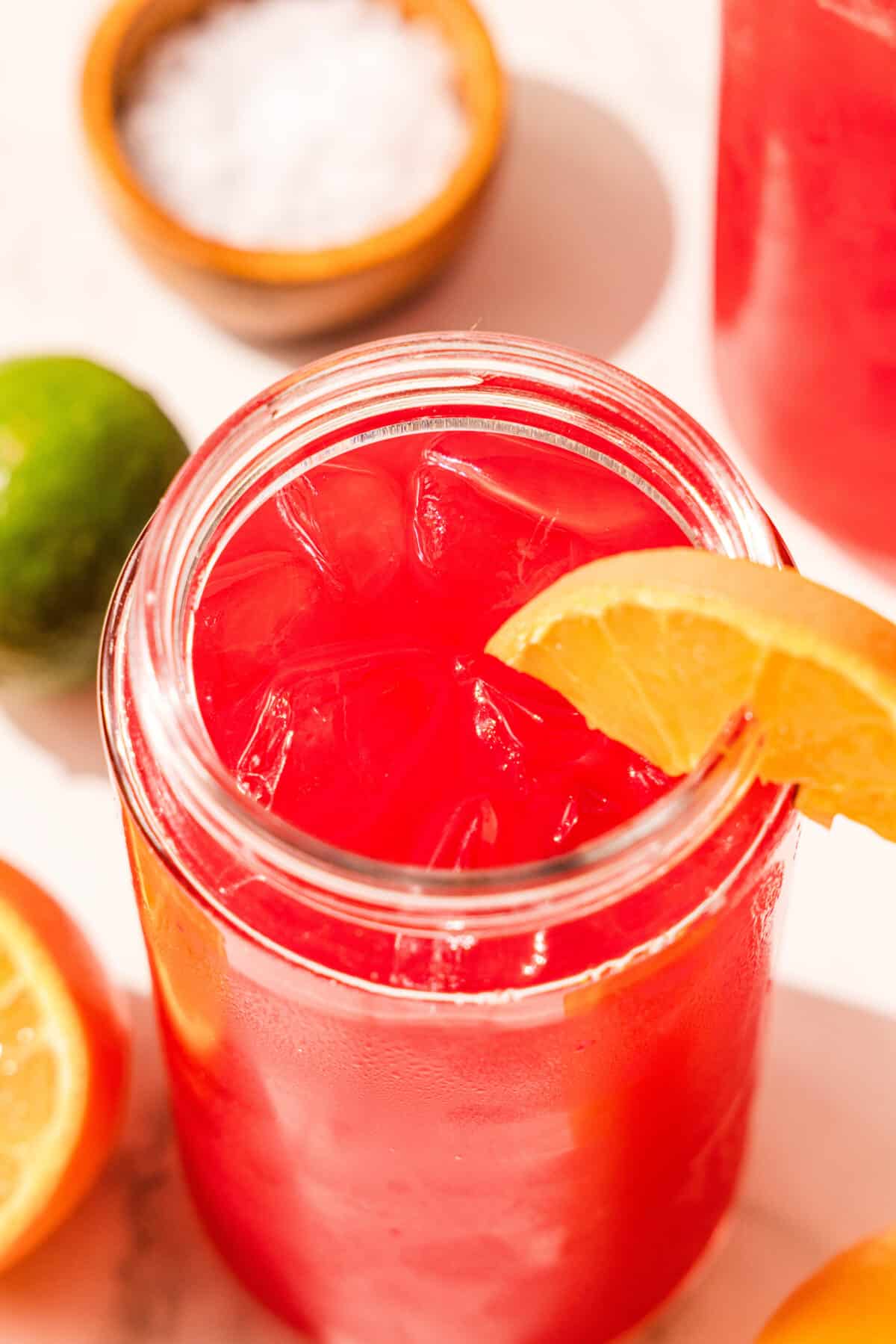 A glass of red juice and ice garnished with an orange slice. Around the glass is a lime, half of an orange, and a bowl of sea salt.