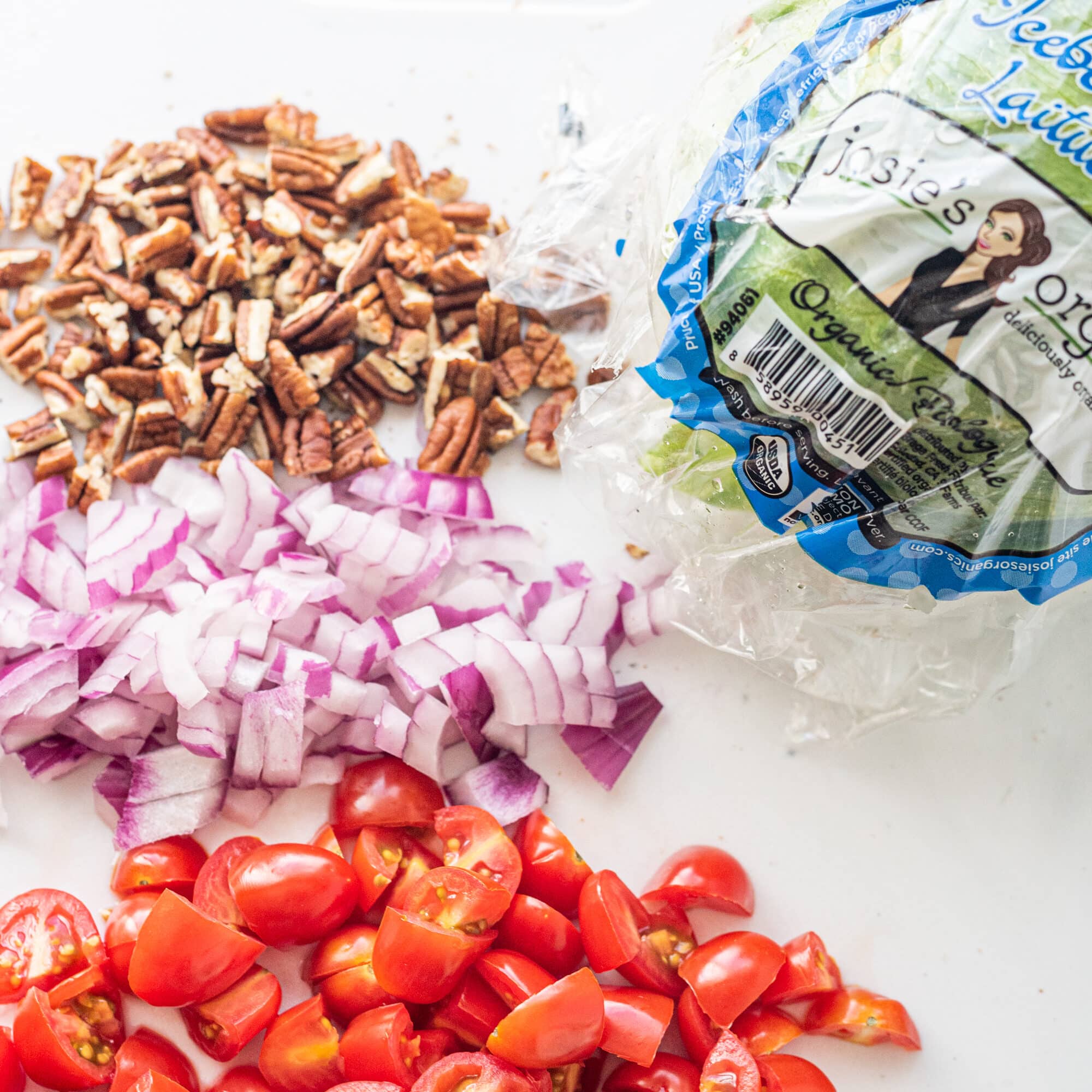 A cutting board with 4 sections of ingredients chopped including pecans, red onion, cherry tomatoes, and iceberg lettuce.