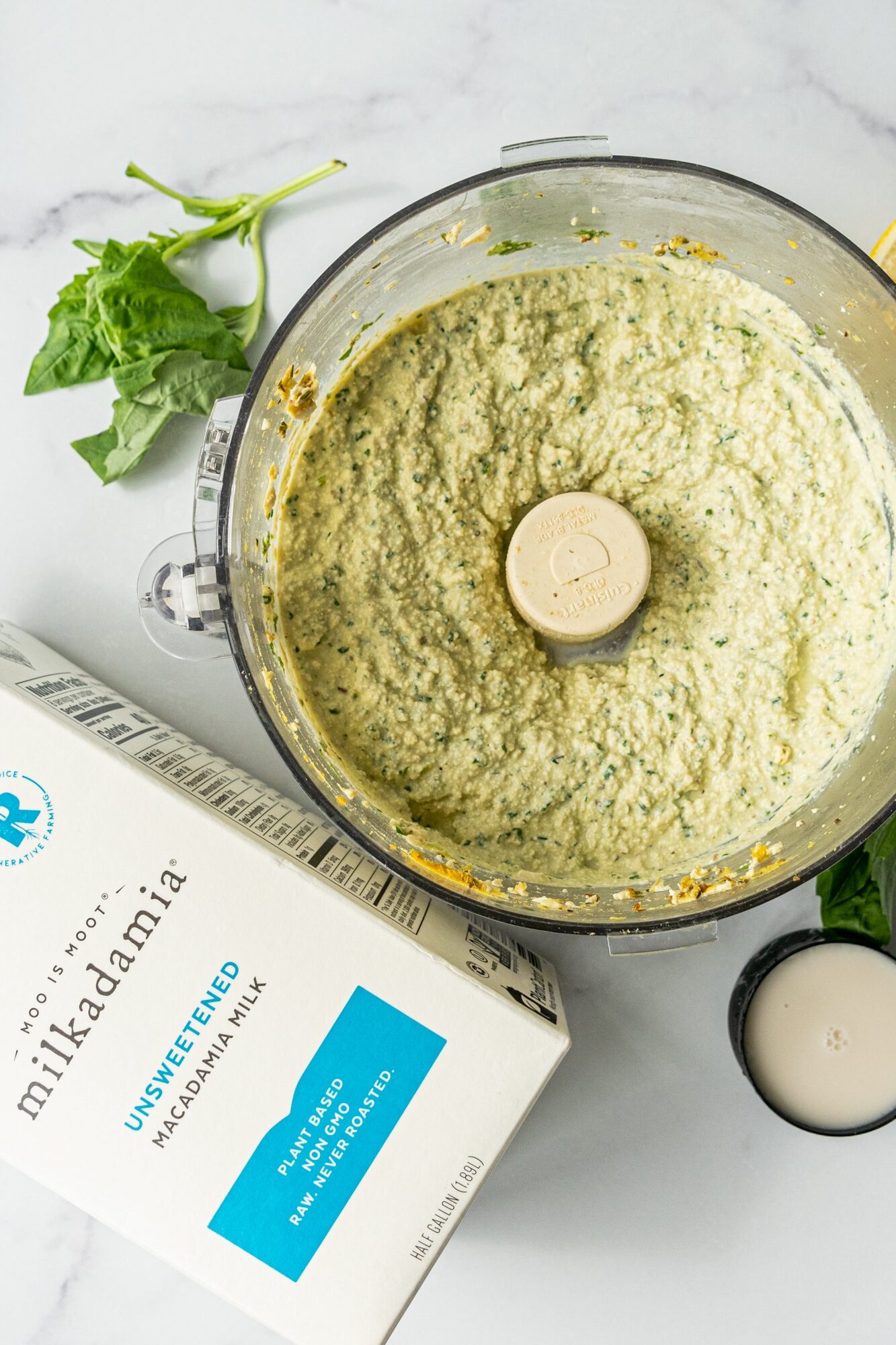 A food processor filled with dairy-free ricotta, a carton of macadamia nut milk, and a sprig of fresh basil.