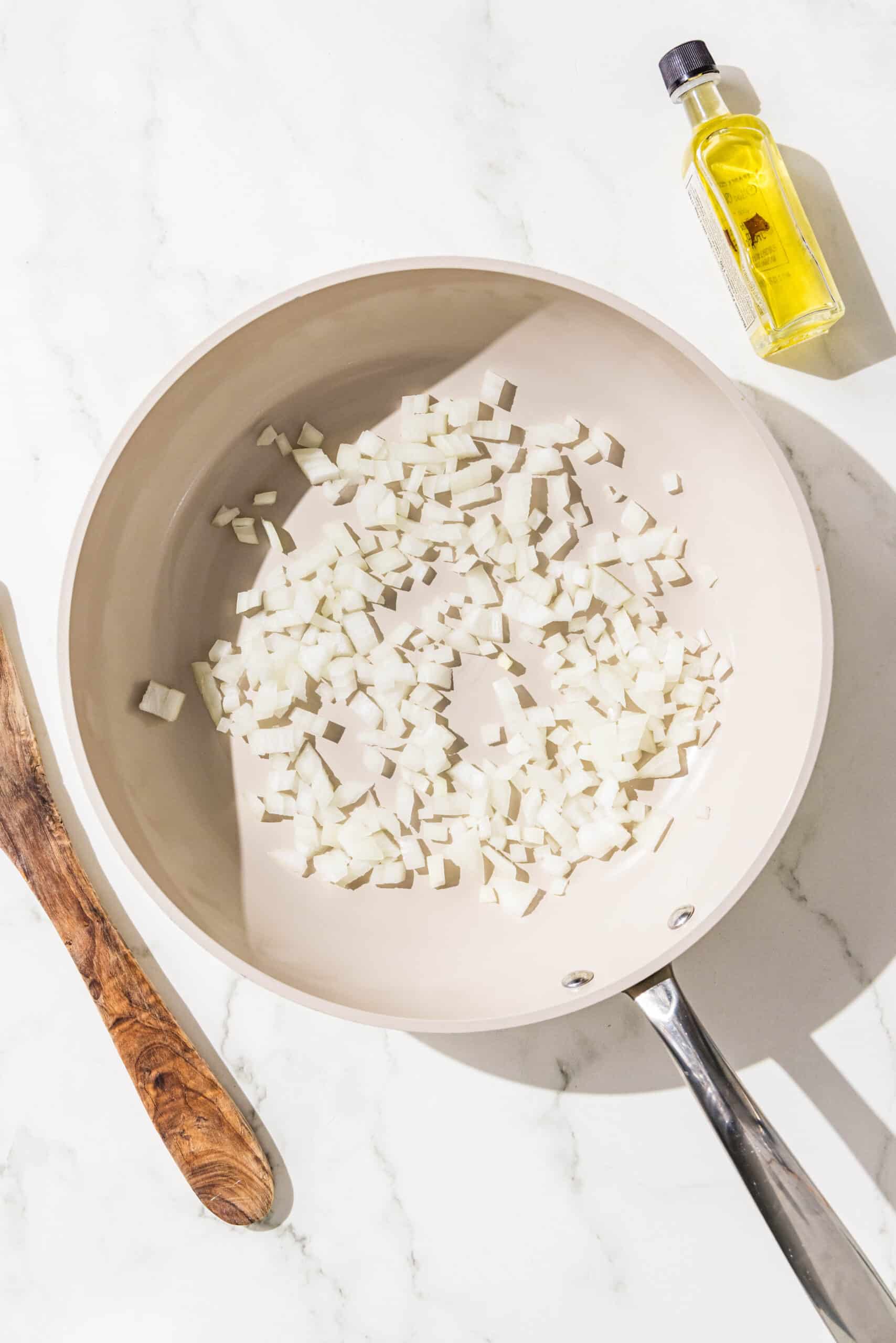 A small white frying pan filled with chopped onion. Beside the pan is a wooden spoon and a small bottle of olive oil.