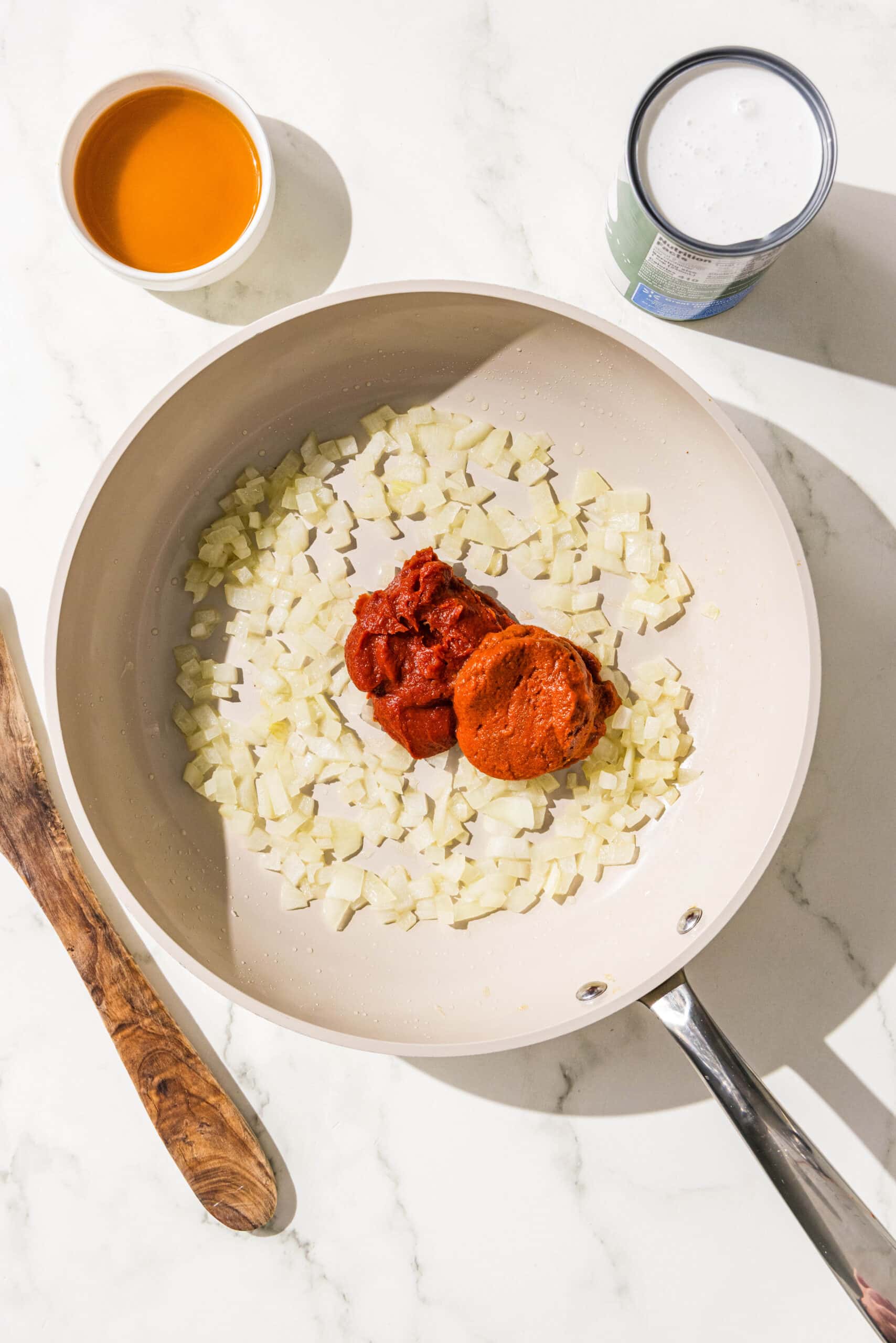 A small frying pan filled with chopped onion and a dollop of tomato paste and red curry paste in the center. Beside the pan is a wooden spoon, a bowl of vegetable broth, and a can of coconut milk.