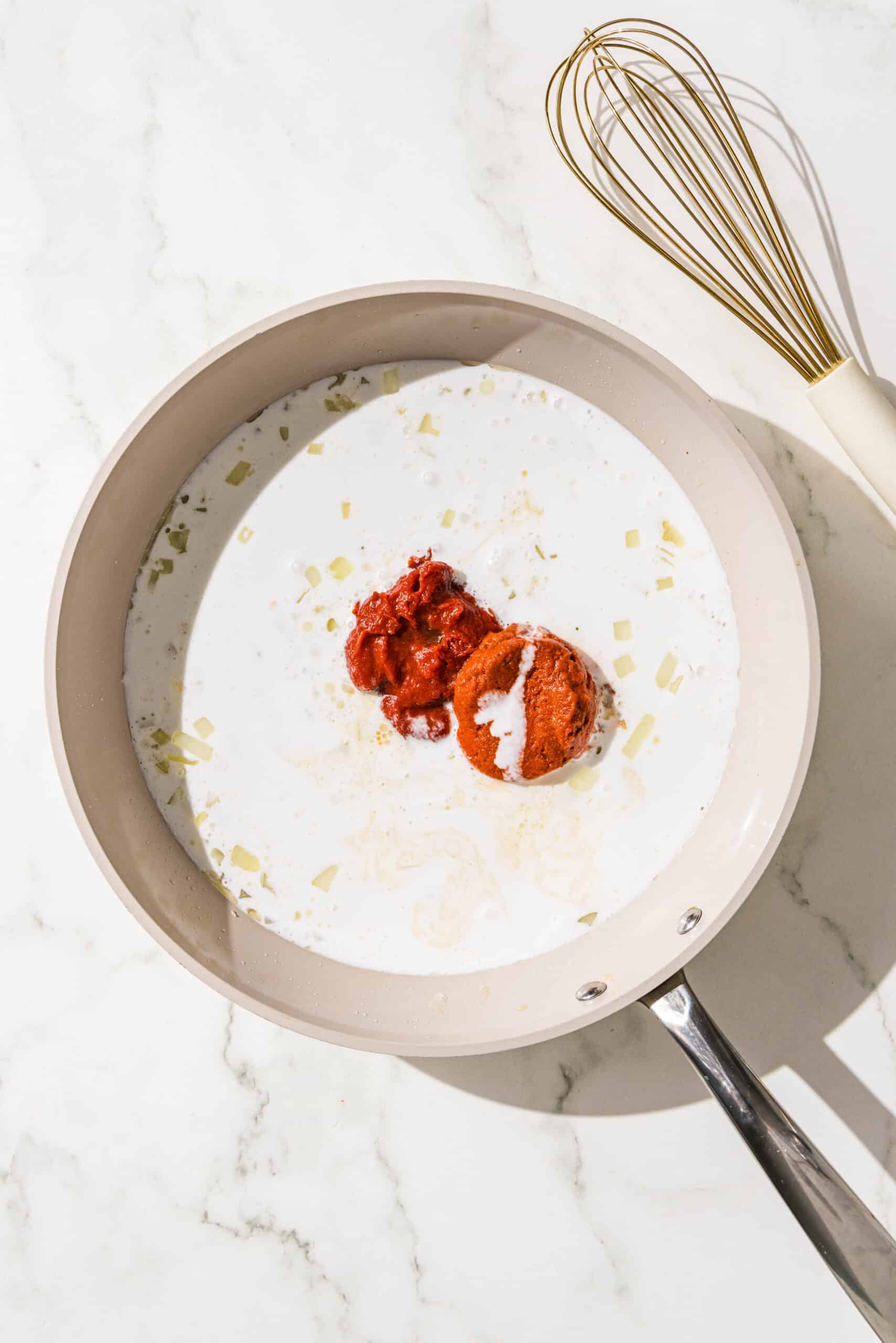 A frying pan filled with coconut milk, red curry paste, tomato paste, and sauteed onions. Beside the pan is a whisk.