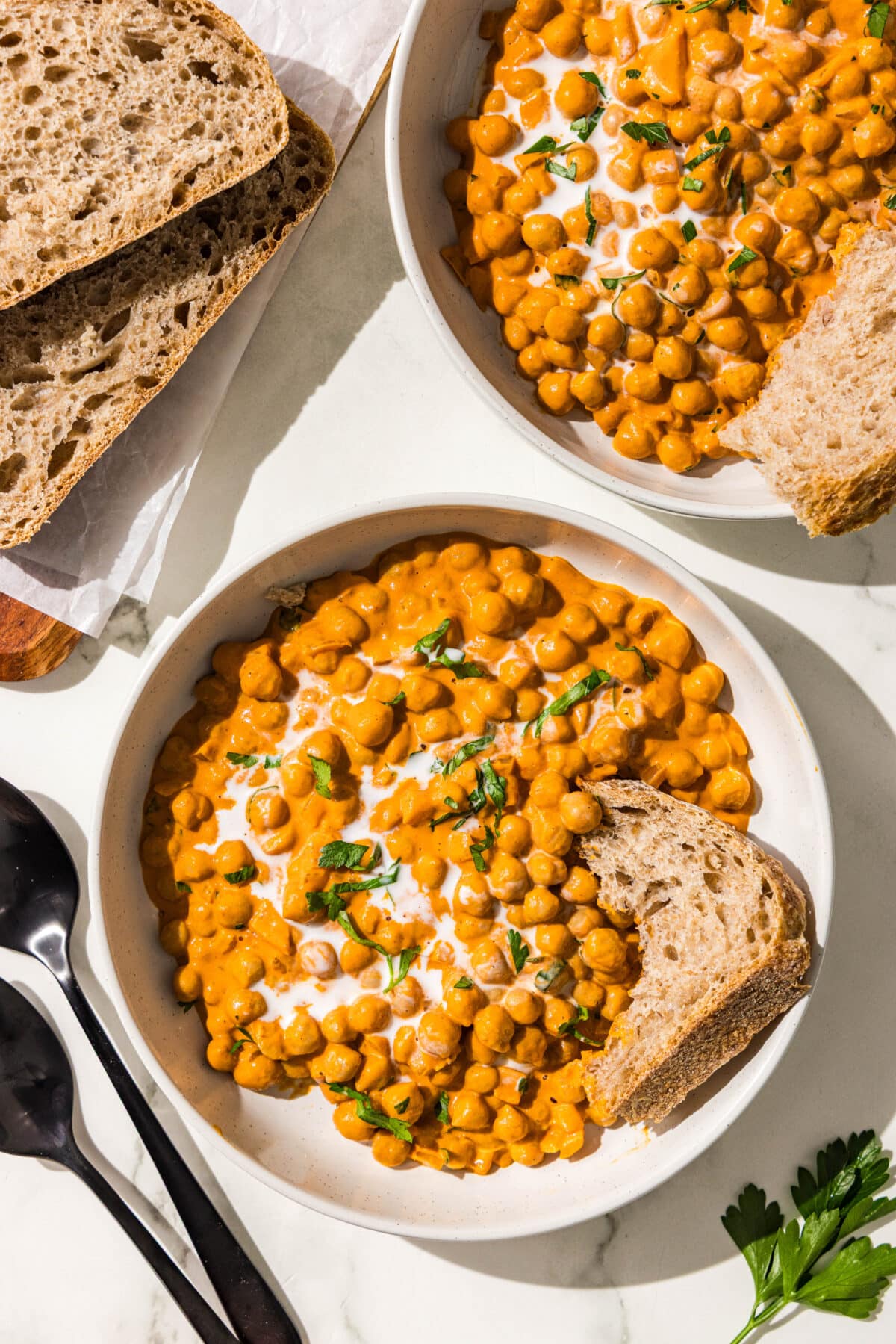 Two bowls of curry chickpeas topped with fresh cilantro, a drizzle of coconut cream, and a piece of bread. Beside the bowls is two pieces of bread and two spoons.
