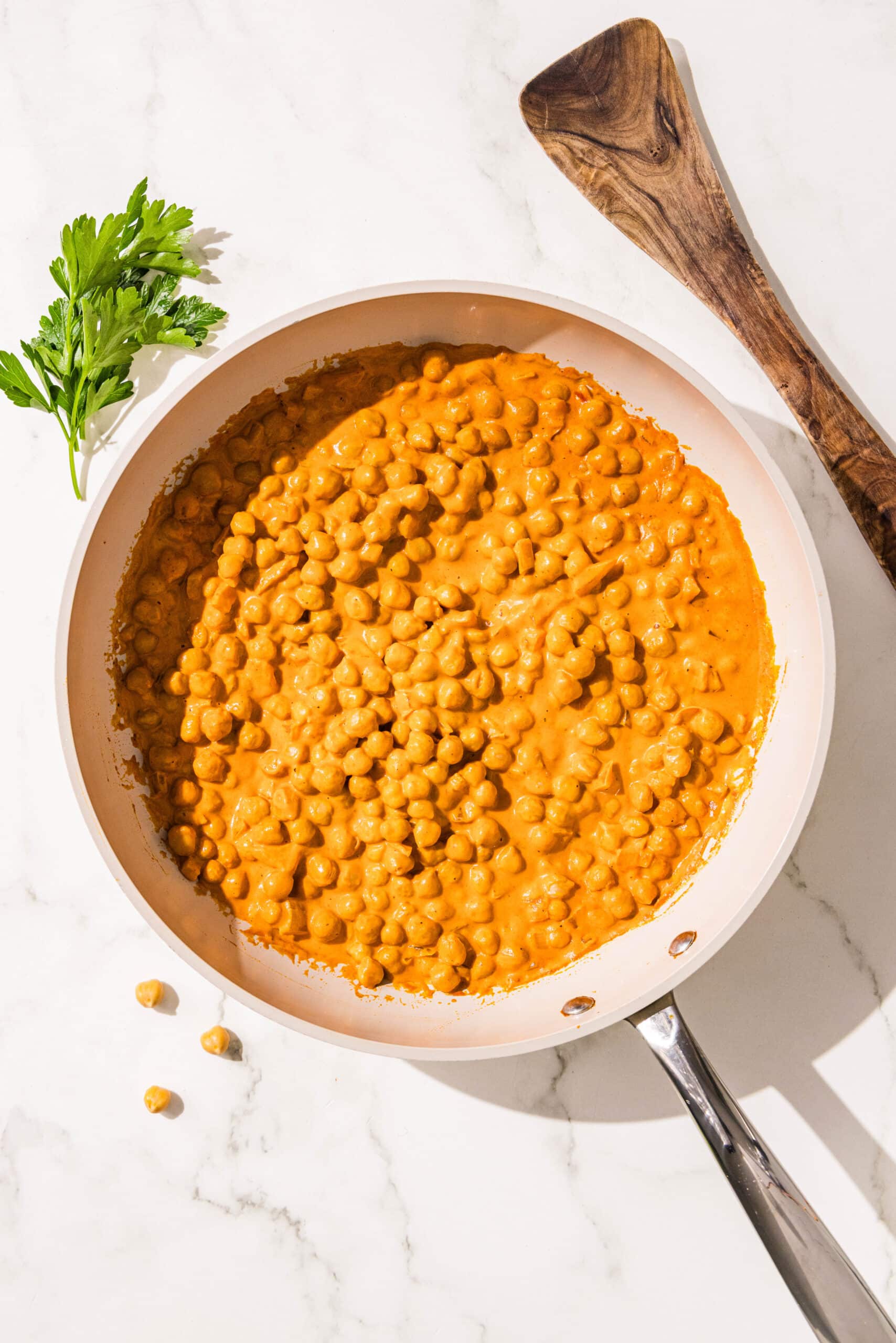 A small frying pan filled with curried chickpeas. Beside the pan is a sprig of fresh cilantro and a wooden spoon.