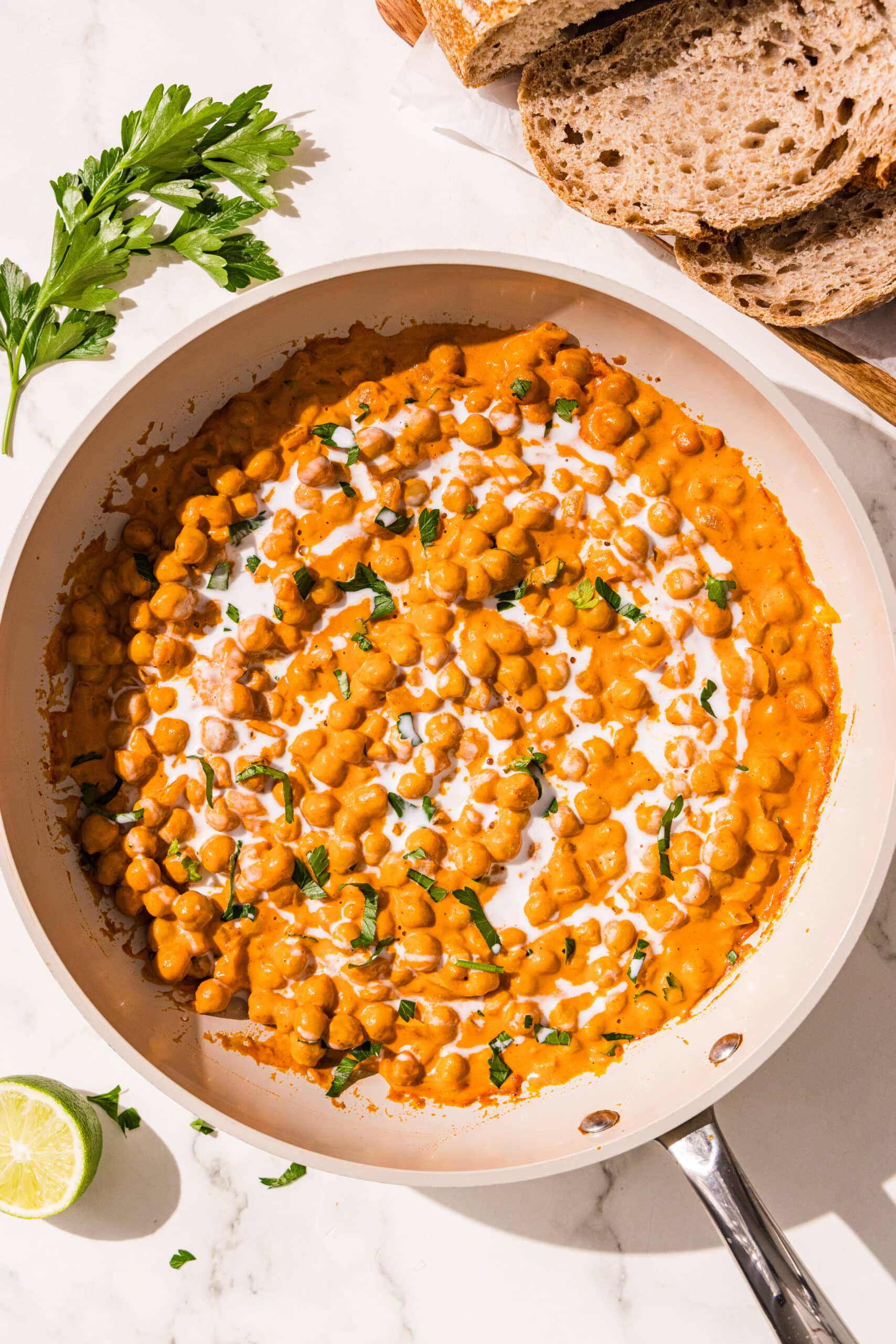 A small frying pan filled with curried chickpeas topped with a drizzle of coconut milk and fresh cilantro. Beside the pan is a sprig of cilantro, three slices of bread, and half of a lime.