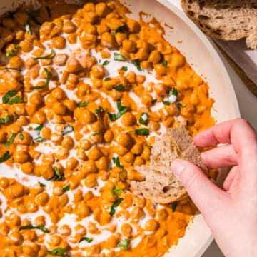 A white pan filled with curry chickpeas topped with fresh cilantro and a drizzle of coconut milk. A white hand is dipping a piece of bread into the chickpeas. Beside the pan are two slices of bread.